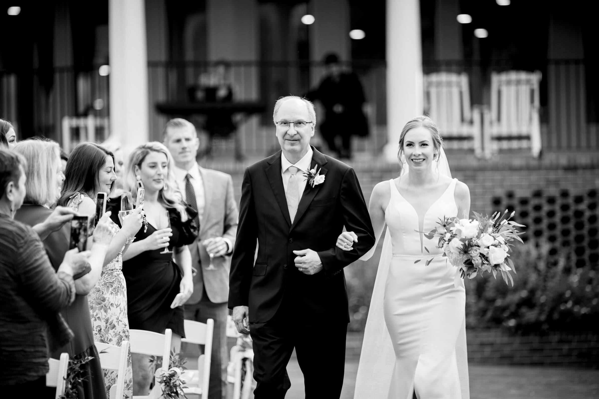 pinehurst wedding photography - lindsey & brendan A bride in a white dress holding a bouquet walks arm-in-arm with an older man in a suit down an aisle. Guests on either side capture the moment with their phones. The setting appears to be an outdoor ceremony with pillars and a patio in the background.