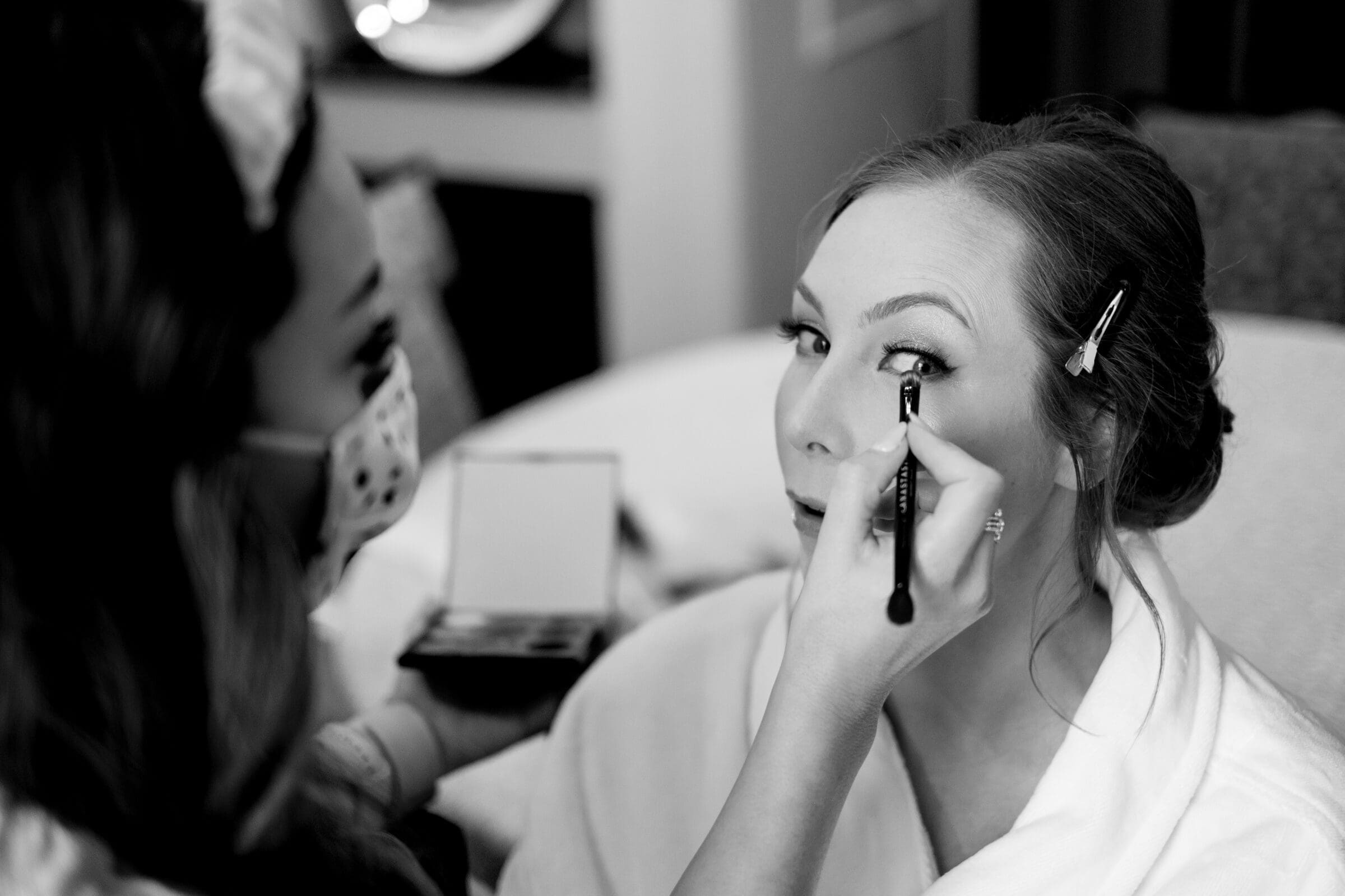 pinehurst wedding photography - lindsey & brendan A woman in a robe sits while a makeup artist applies eyeliner. The artist holds an eyeliner pencil and a palette, focused on the task. The setting is calm and intimate, capturing the moment in black and white.