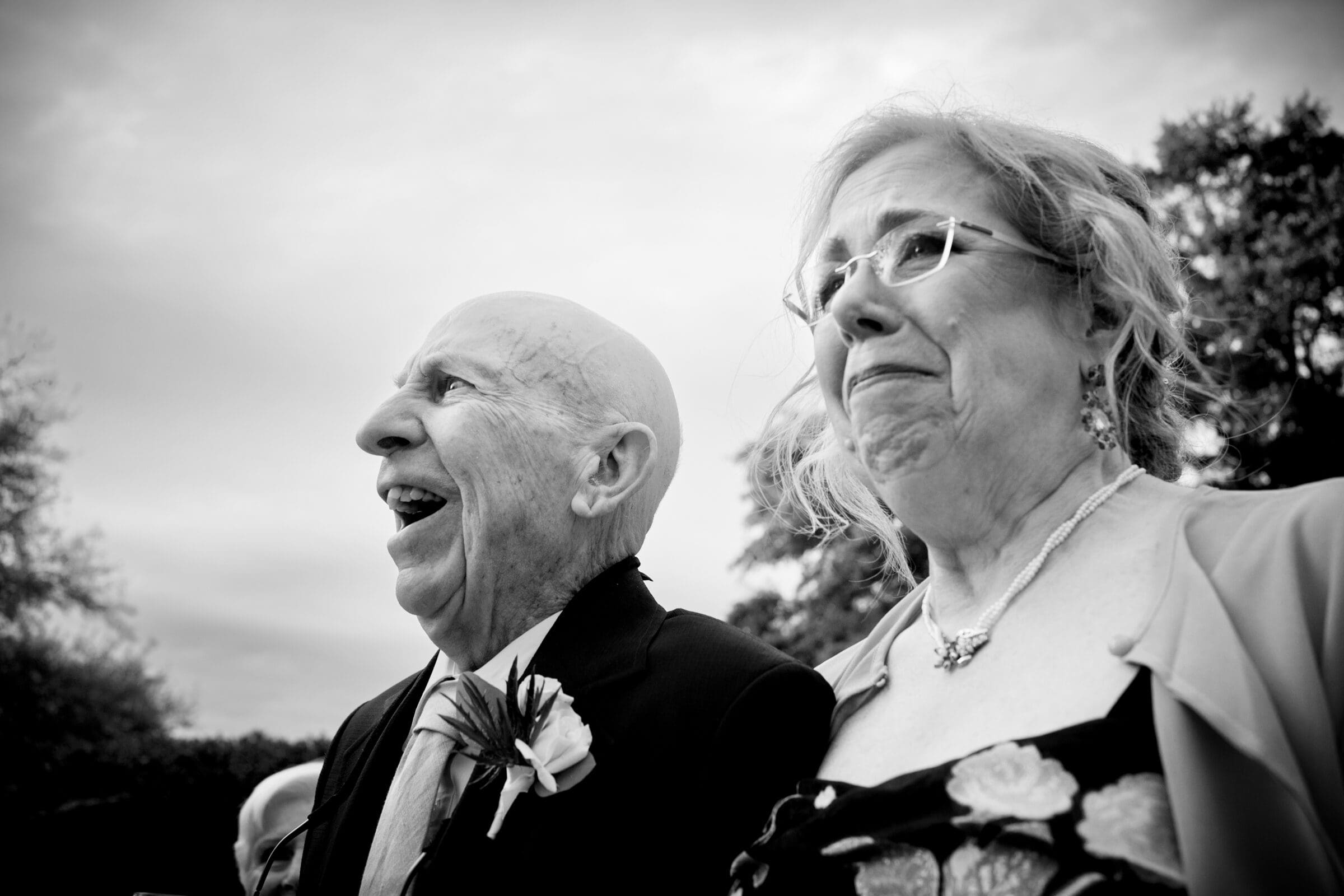 pinehurst wedding photography - lindsey & brendan An elderly couple is standing outside, smiling and looking emotional. The man is laughing heartily, wearing a suit with a boutonniere, while the woman has a joyful expression, dressed in a floral outfit. Trees are visible in the background.