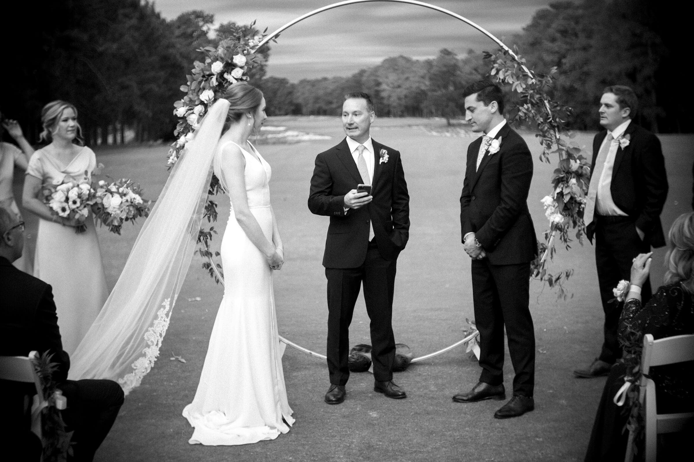 pinehurst wedding photography - lindsey & brendan A black-and-white photo of an outdoor wedding ceremony. A bride and groom stand facing each other under a floral arch, accompanied by the officiant. Bridesmaids and groomsmen are present. The background features trees and a cloudy sky.