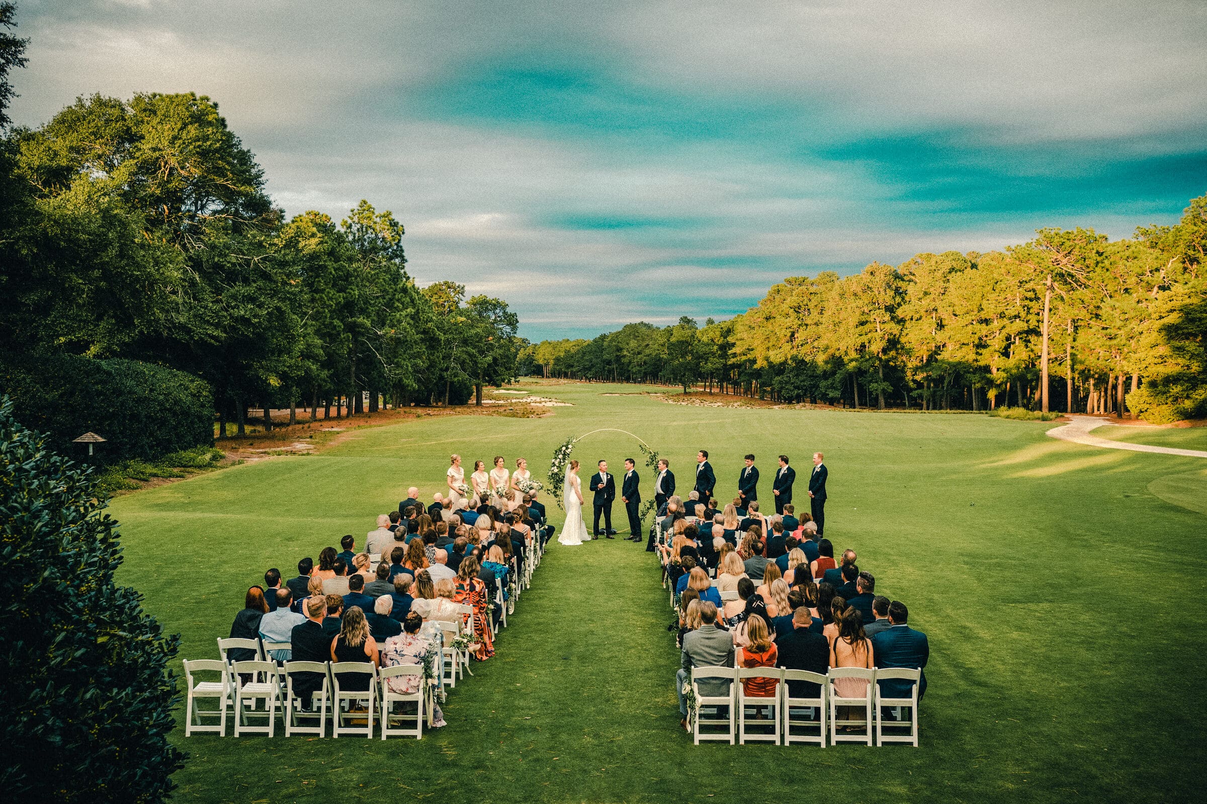 An outdoor wedding ceremony on a green field surrounded by trees. Guests are seated in rows facing a couple under a white arch. The sky is partly cloudy, casting a soft light over the scene.