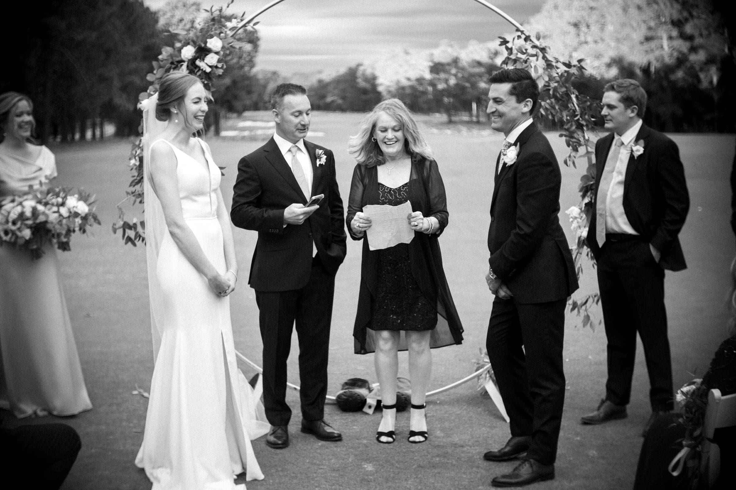 pinehurst wedding photography - lindsey & brendan A black and white image of a wedding ceremony outdoors. The bride and groom are smiling, standing under a circular floral arch. The officiant is holding a script, flanked by bridesmaids and groomsmen dressed in formal attire.