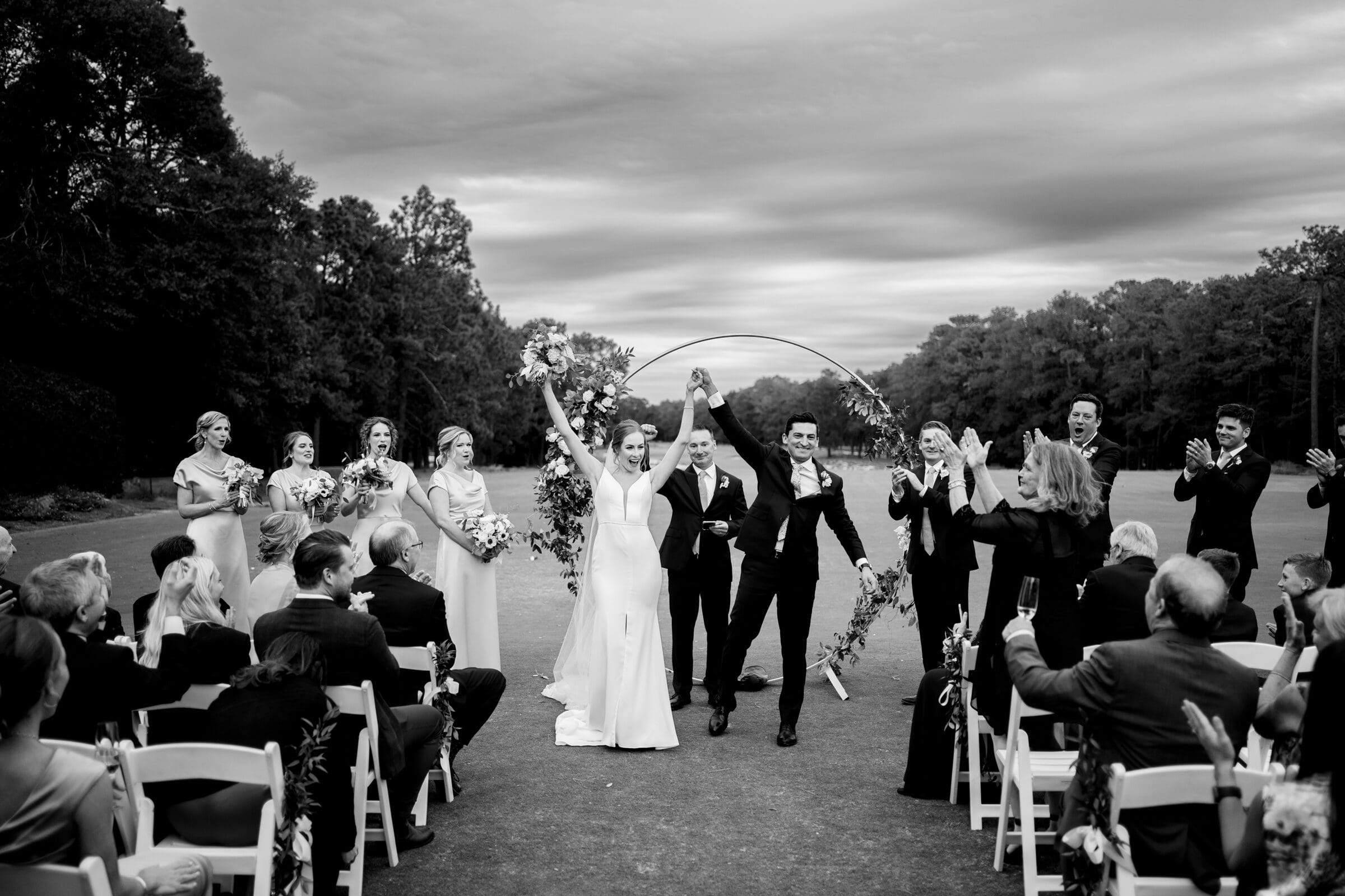 A joyful bride and groom walk arm in arm down the aisle, celebrating with raised hands. They are surrounded by cheering bridesmaids and groomsmen. Guests clap in a scenic outdoor setting under a dramatic cloudy sky at Pinehurst.