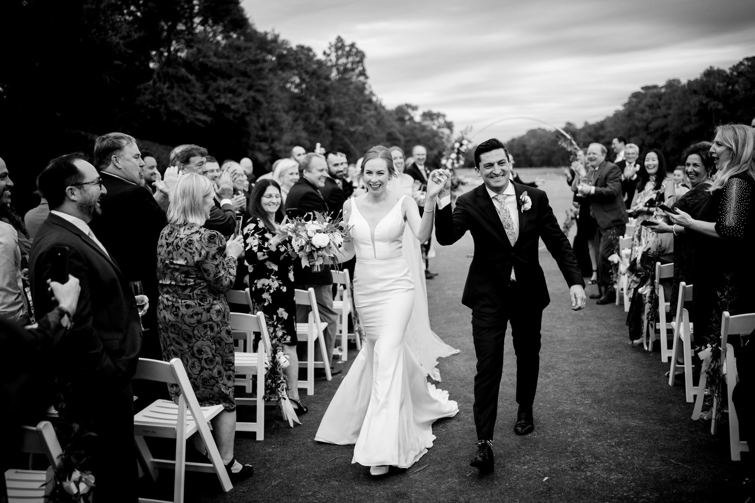 pinehurst wedding photography - lindsey & brendan A newlywed couple joyfully walks down the aisle, surrounded by smiling guests on either side. The bride holds a bouquet, and both are dressed in formal wedding attire. The setting is outdoors with trees in the background. Black and white image.