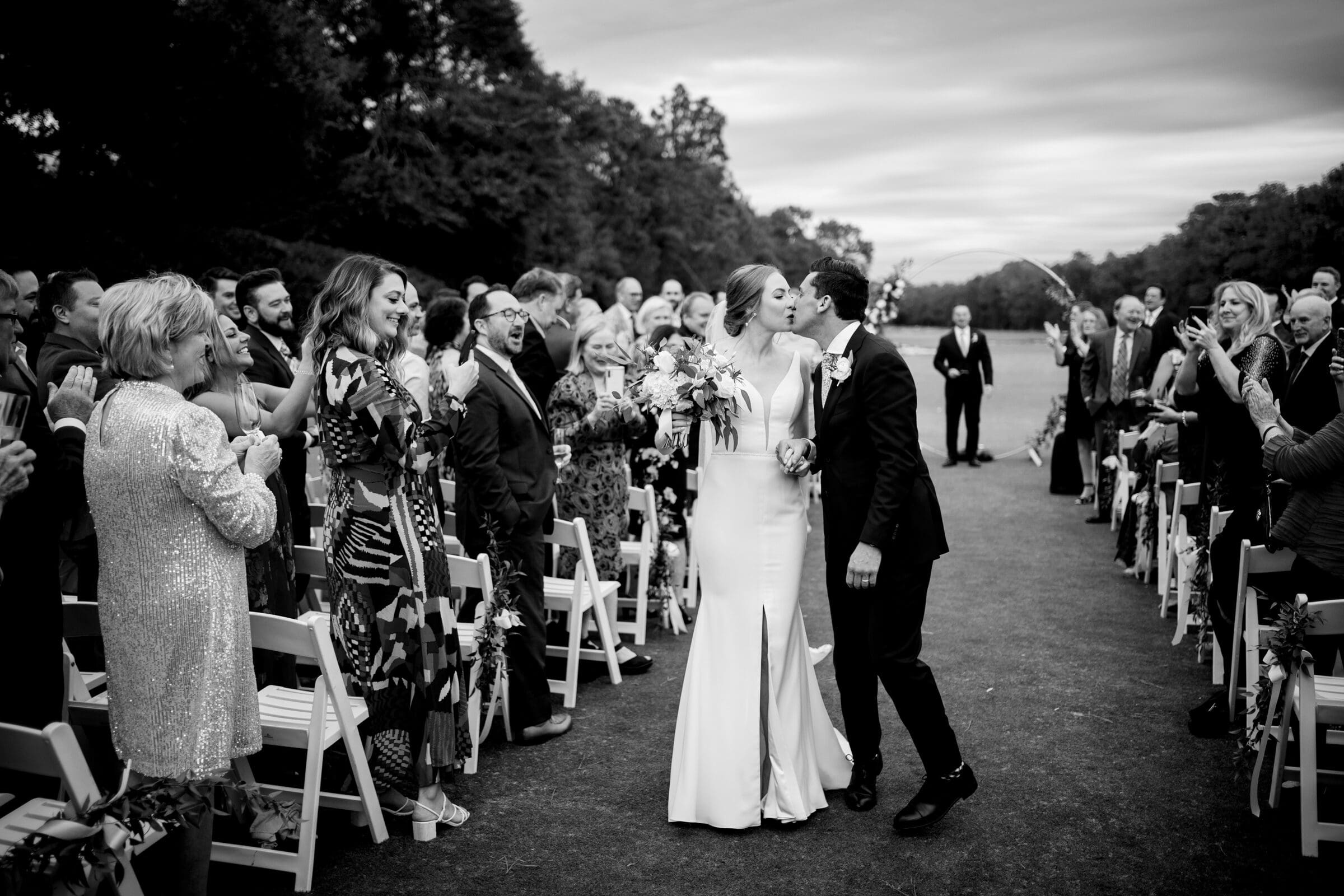 pinehurst wedding photography - lindsey & brendan A black and white photo of a newlywed couple kissing while walking down an outdoor aisle lined with cheering guests. The bride holds a bouquet, and the groom wears a suit. White chairs and trees are in the background.