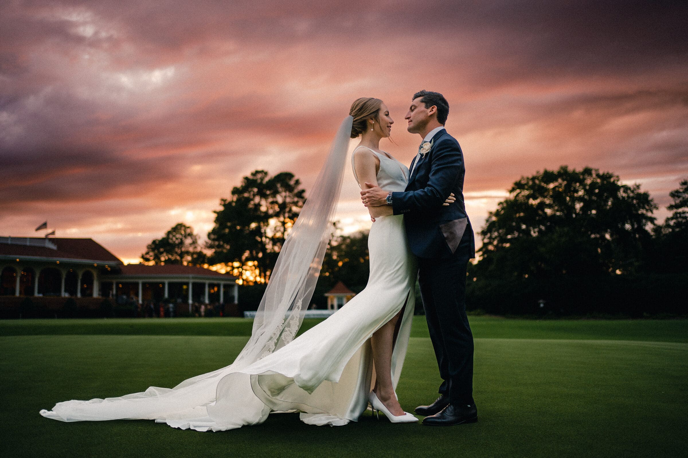 pinehurst wedding photography - lindsey & brendan A bride and groom embrace on a lawn at sunset. The bride wears a long white gown and veil, and the groom is in a dark suit. The sky is a dramatic mix of orange and purple hues, and trees silhouette the background.