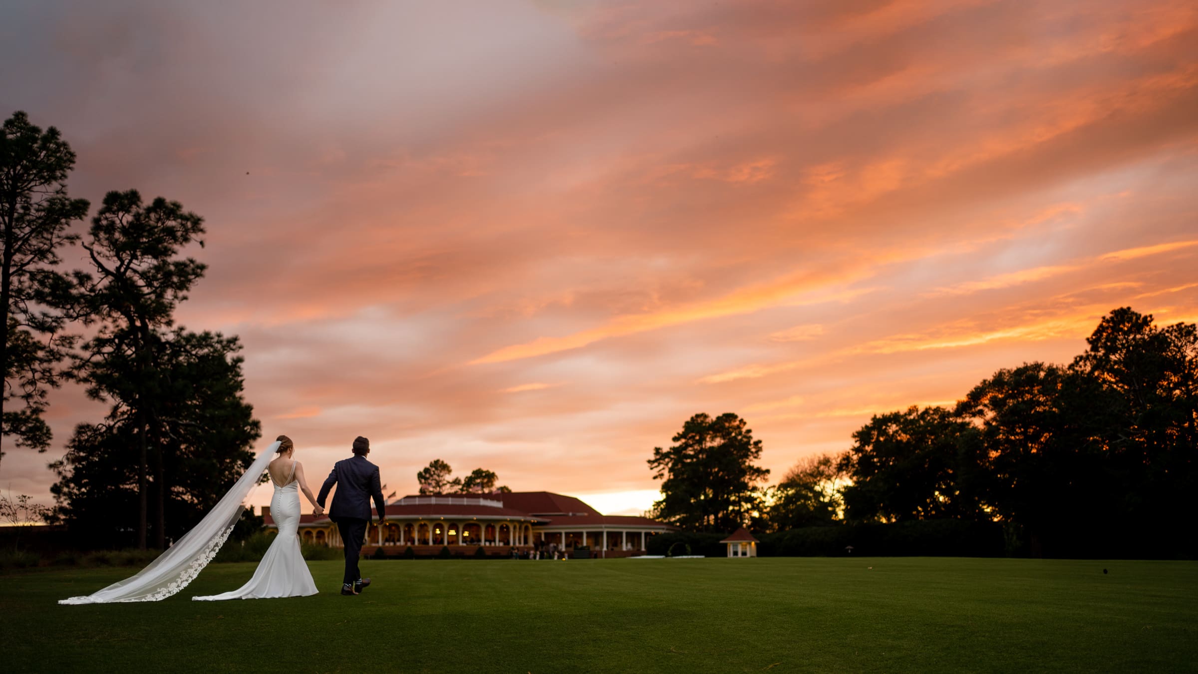 A bride and groom walk hand in hand across a manicured lawn at sunset. The sky is filled with orange and pink hues, and a grand building with arches is visible in the background. Trees frame the scene, creating a romantic atmosphere.