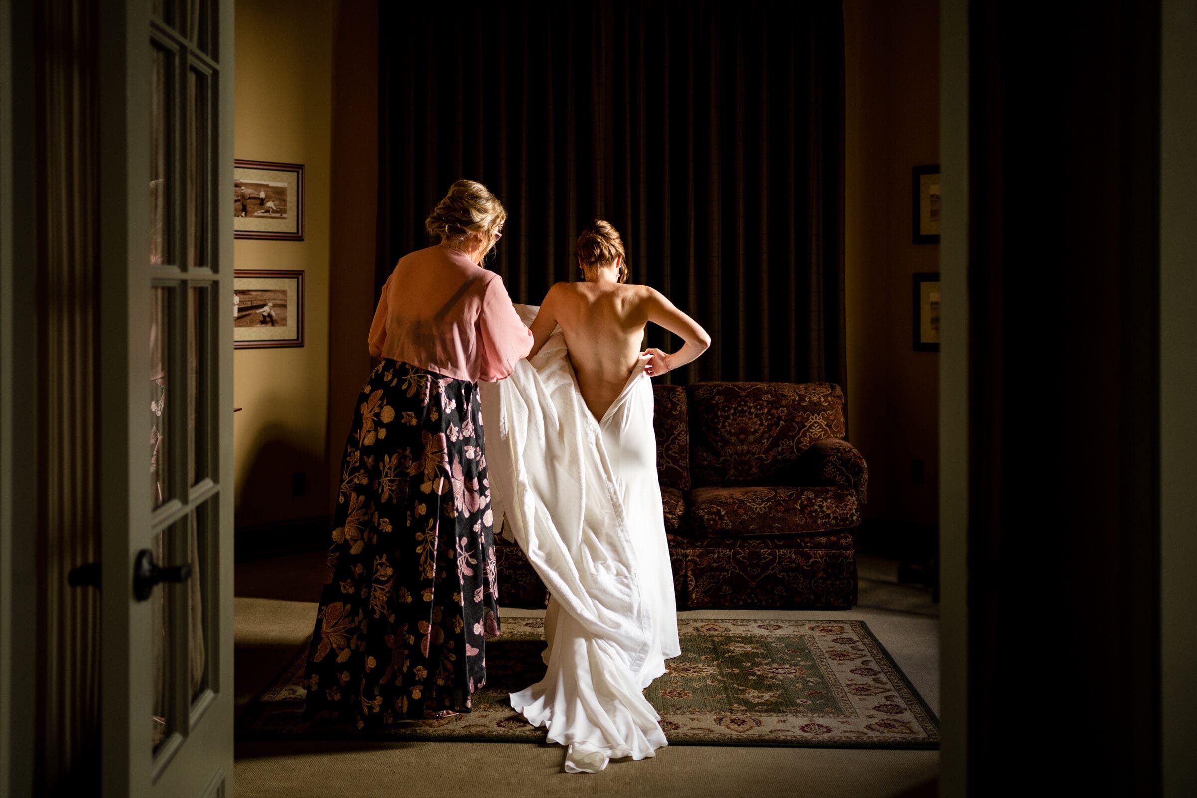 pinehurst wedding photography - lindsey & brendan A bride in a white gown stands in a warmly lit room, adjusting her dress with the help of a woman in a floral dress. They are near a rug and an armchair, with framed pictures on the walls, under draped curtains.