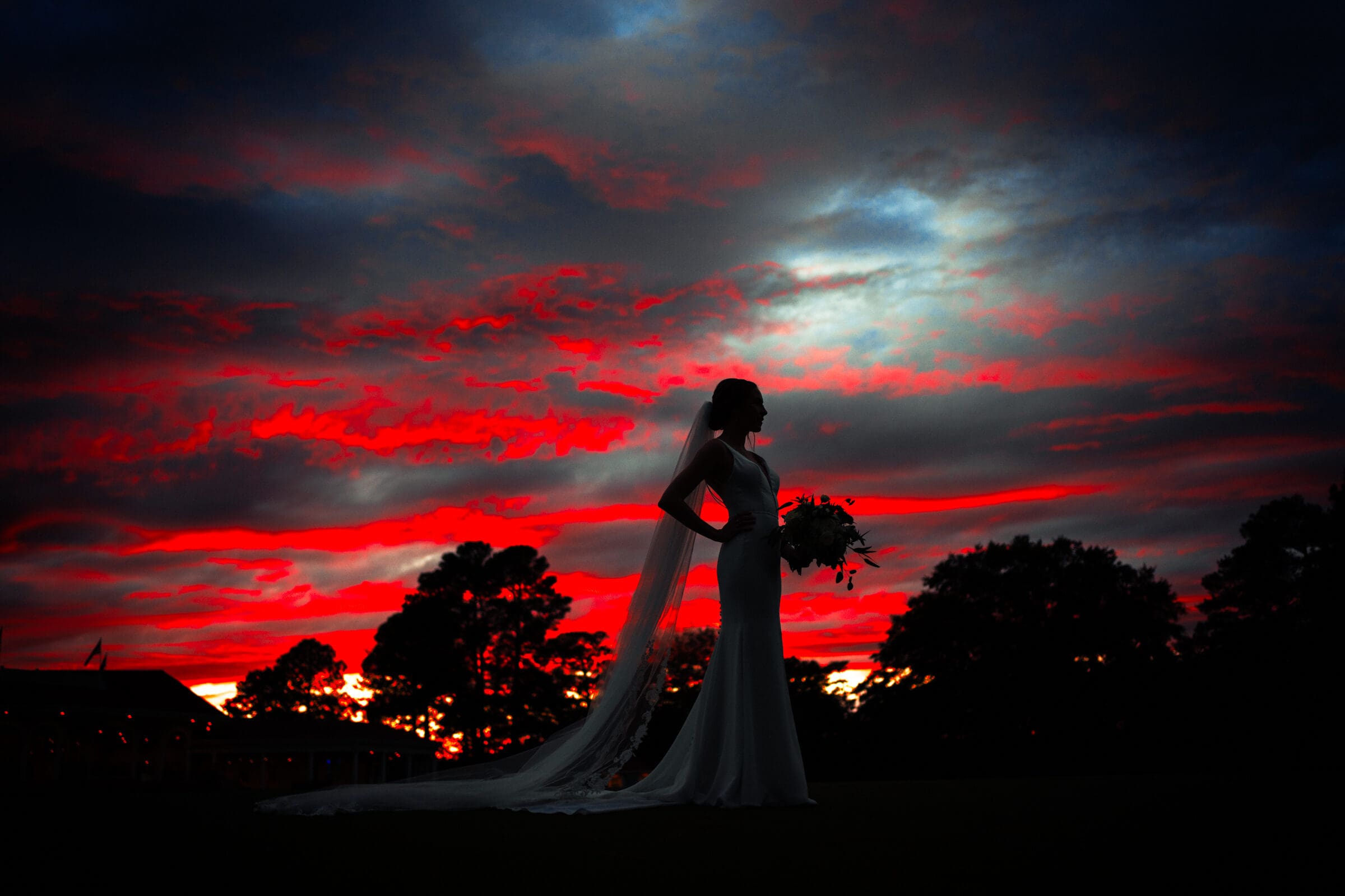 pinehurst wedding photography - lindsey & brendan A silhouetted bride holding a bouquet stands against a dramatic sky at sunset, with vibrant red and blue clouds. Her veil and dress trail elegantly behind her, creating an ethereal scene. Trees and a building are faintly visible on the horizon.