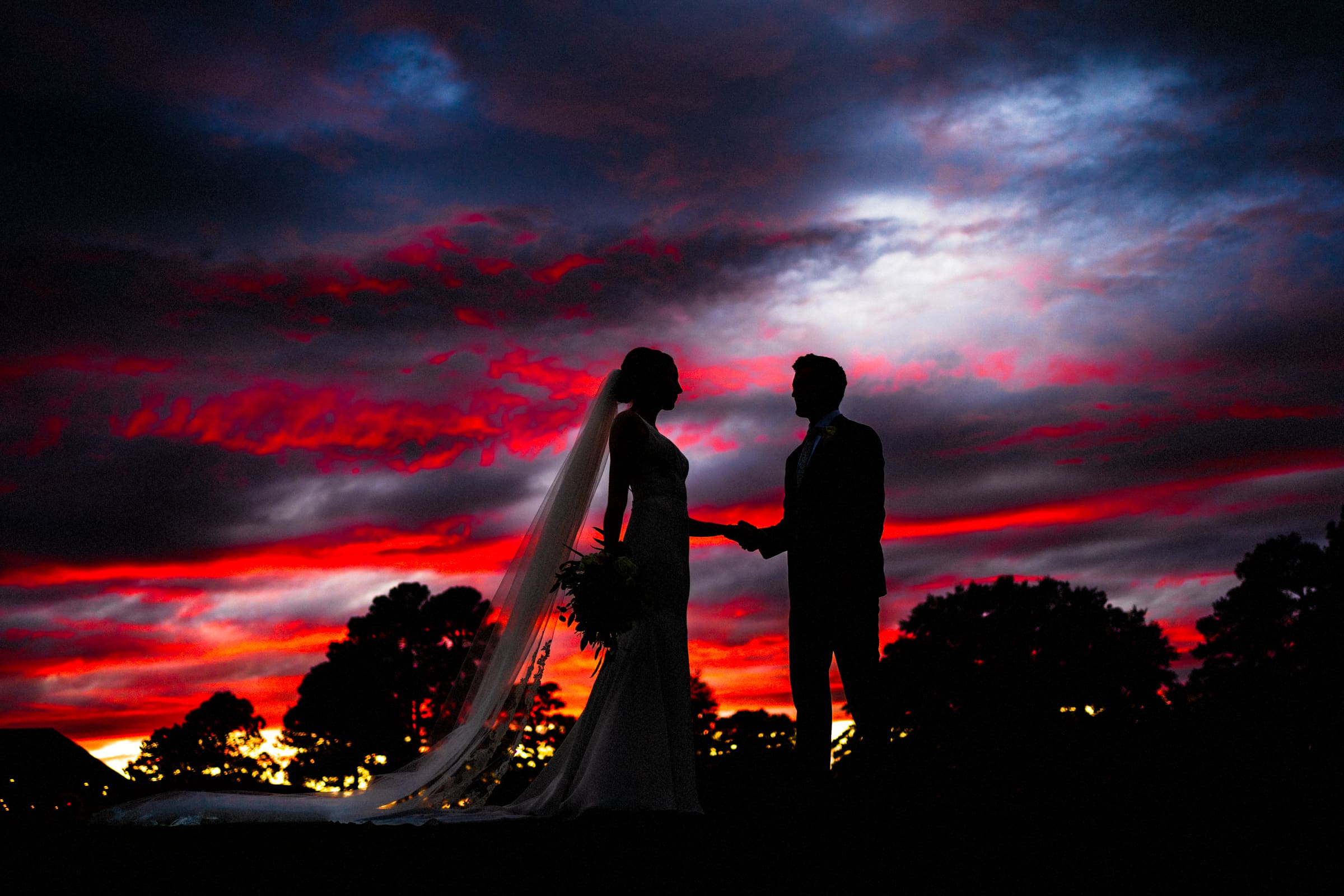Silhouetted bride and groom hold hands against a dramatic sunset sky painted in fiery reds and deep blues, with trees lining the horizon.