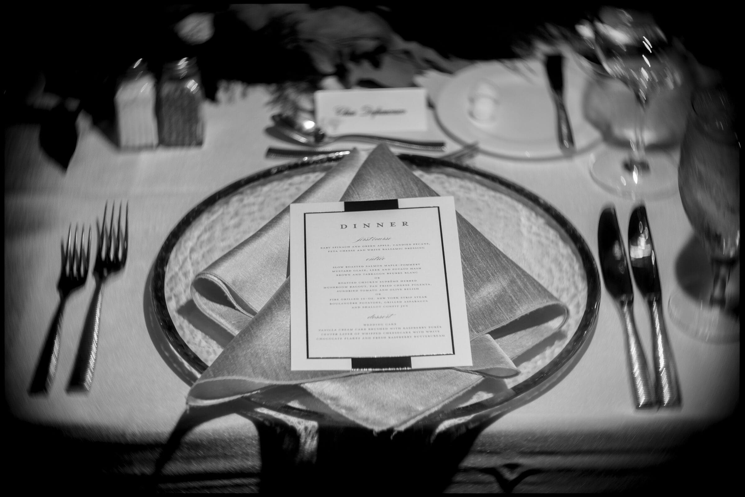 pinehurst wedding photography - lindsey & brendan Black and white image of an elegant dinner table setting. A menu is centered on a folded napkin placed on a plate. Silverware is arranged neatly on either side, with glassware above. The ambiance is formal, suggesting a fine dining experience.