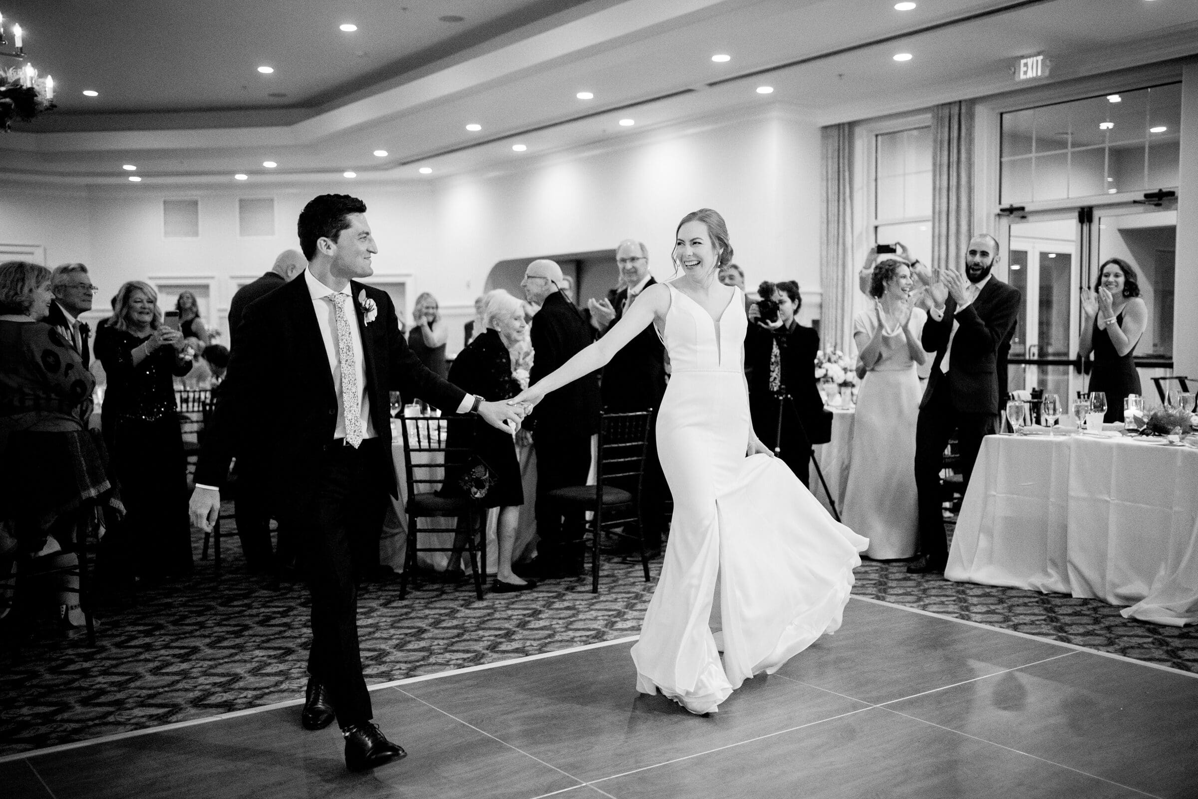 pinehurst wedding photography - lindsey & brendan A bride and groom share a joyful dance at their wedding reception. The groom wears a suit, and the bride is in a flowing gown. Guests in formal attire watch and applaud in a warmly lit hall.