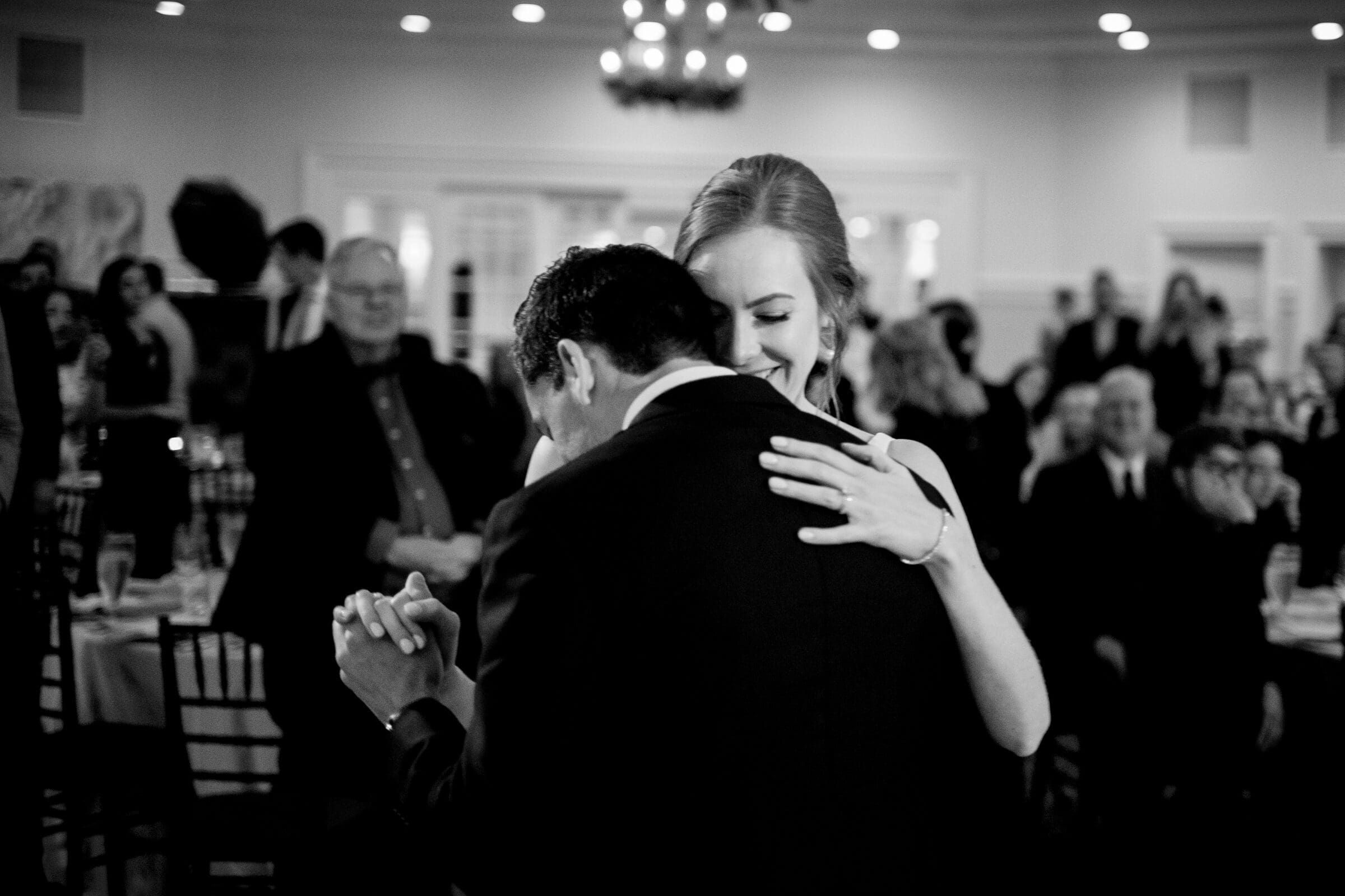 pinehurst wedding photography - lindsey & brendan A black and white photo of a couple embracing and dancing in a room full of guests. The woman is smiling with her eyes closed. The guests are seated at tables, blurred in the background, giving a sense of a formal event.