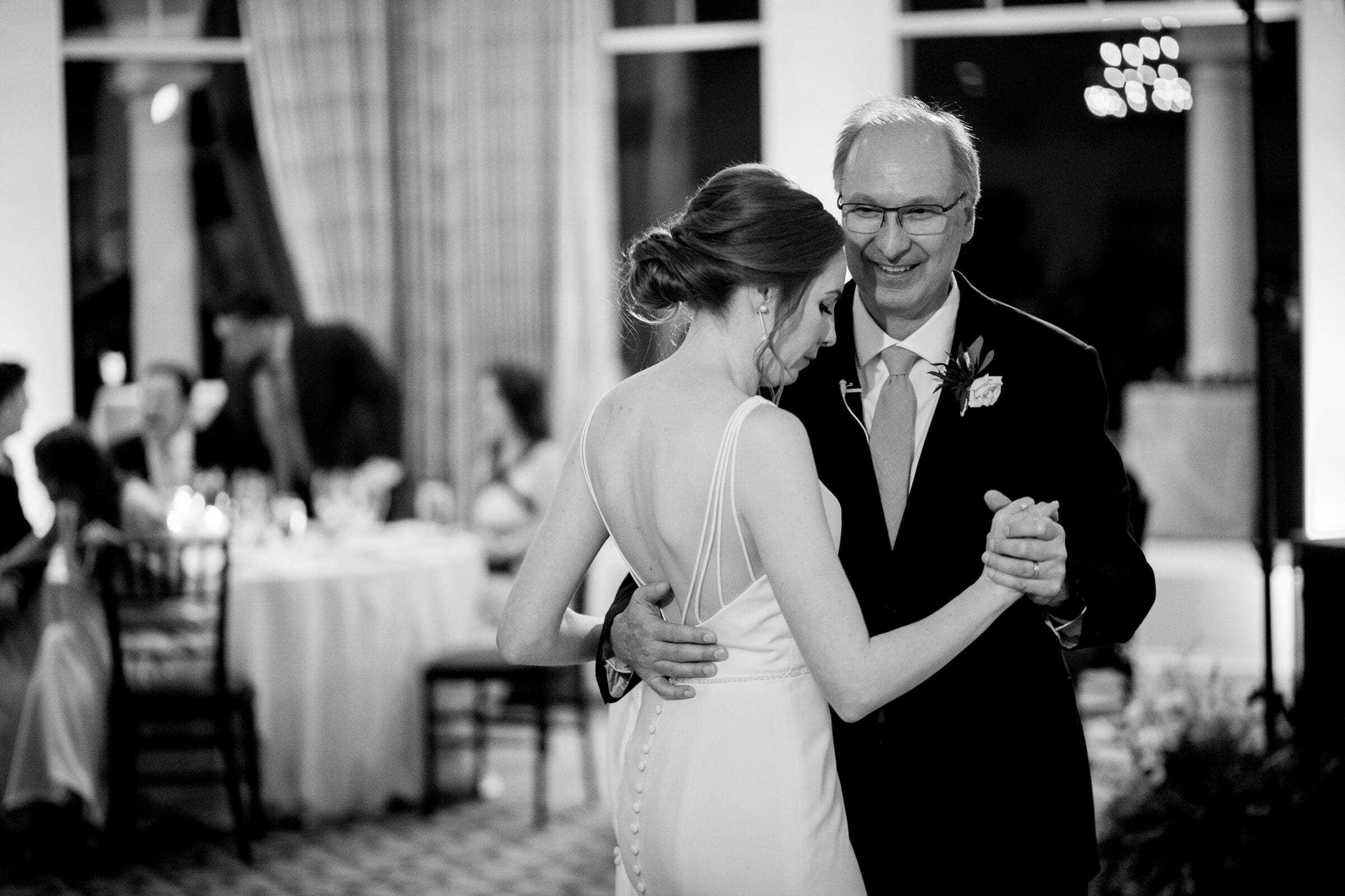 pinehurst wedding photography - lindsey & brendan A black-and-white photo of a smiling couple dancing. The woman is in a white dress with her hair up, and the man is in a suit and tie with glasses. They are in a warmly lit room with guests seated at tables in the background.