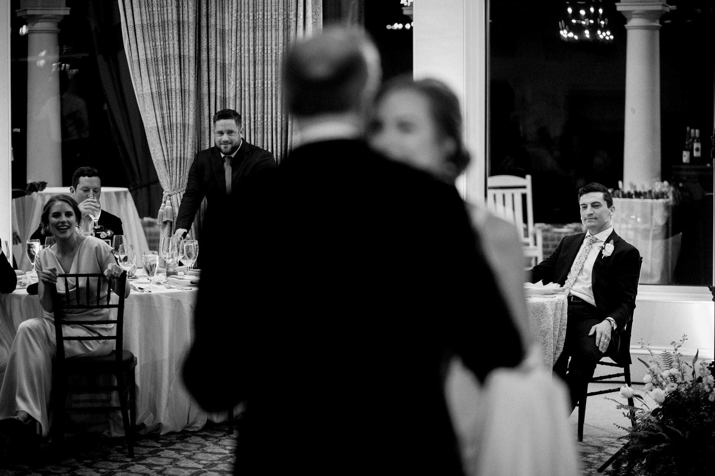 pinehurst wedding photography - lindsey & brendan A black and white image of a wedding reception. A couple dances in the foreground, slightly blurred. Guests sit at round tables watching. One man, seated separately, gazes at the dancing couple. The room is elegantly decorated with large windows.