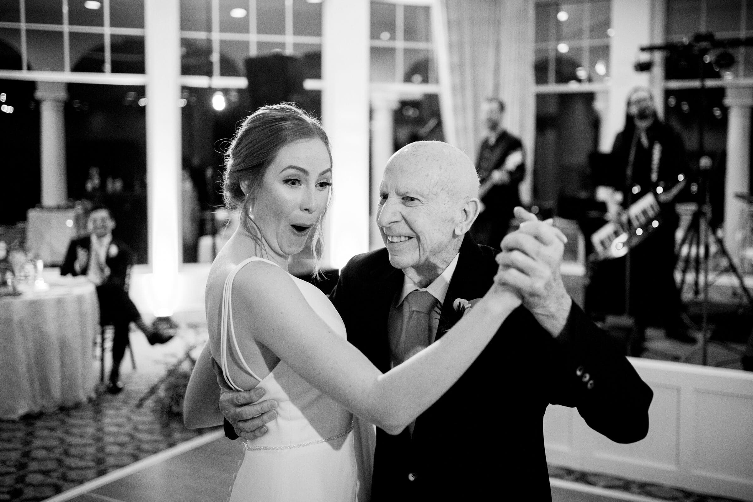 pinehurst wedding photography - lindsey & brendan A black and white photo of a young woman in a white dress dancing joyfully with an elderly man in a suit. They are smiling, and there is a band performing in the background.