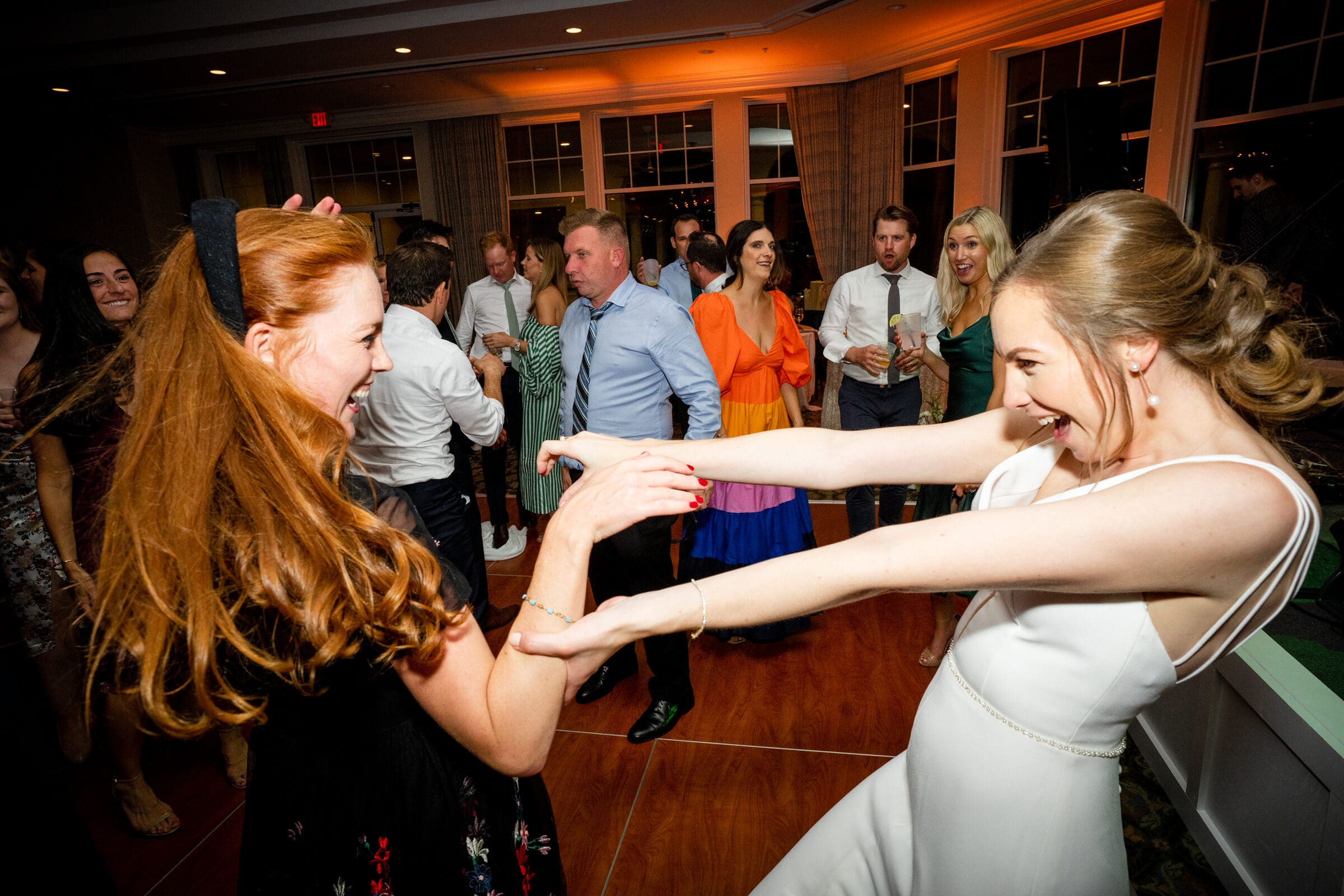 pinehurst wedding photography - lindsey & brendan Two women smiling and dancing energetically at a party. One wears a white dress, and the other wears a black dress with long red hair. People in colorful outfits are in the background, under warm lighting.