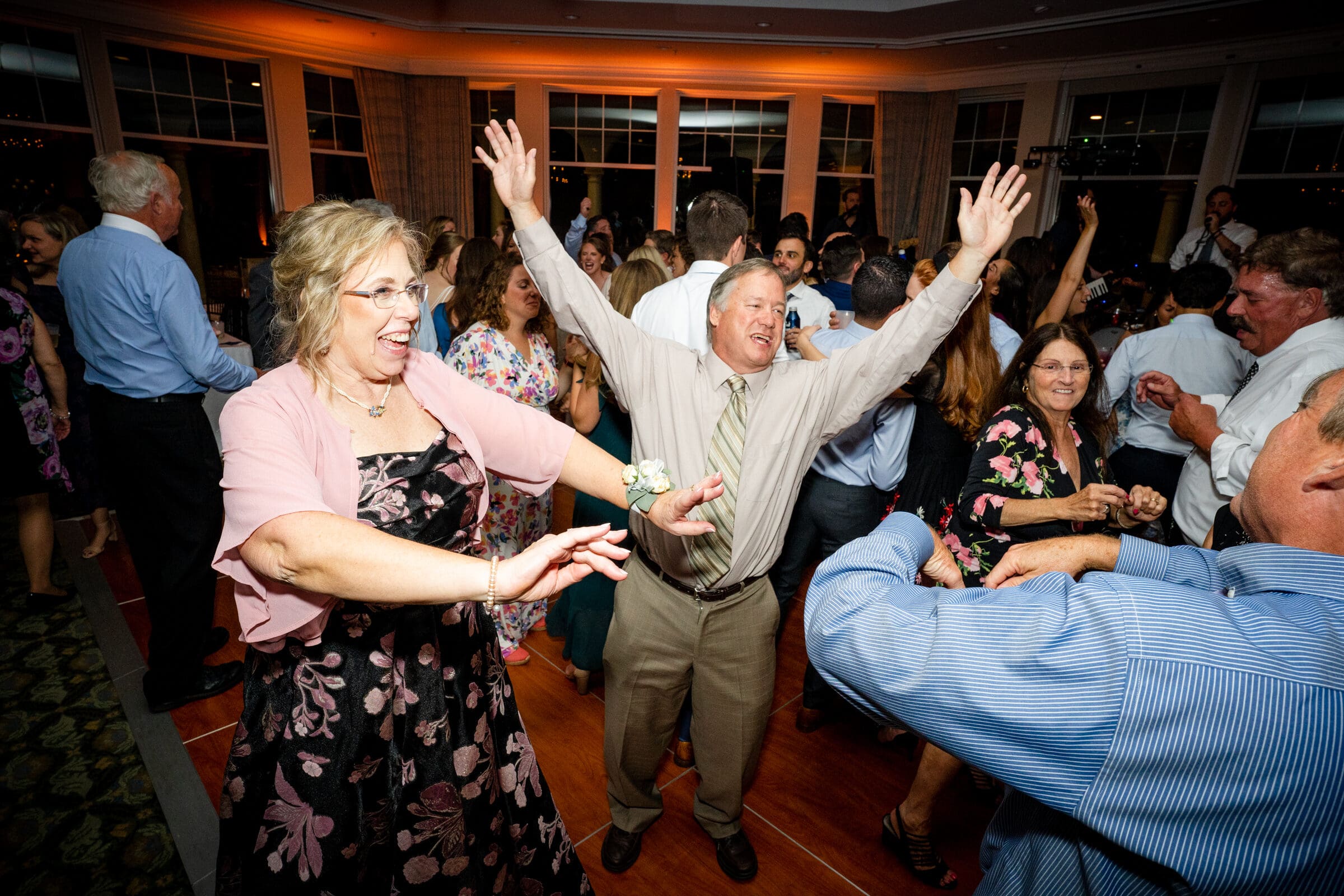 pinehurst wedding photography - lindsey & brendan A joyful crowd dancing at a party, with a man in a light shirt and tie energetically raising his arms and a woman in a floral dress dancing nearby. The room is filled with people enjoying the festive atmosphere.