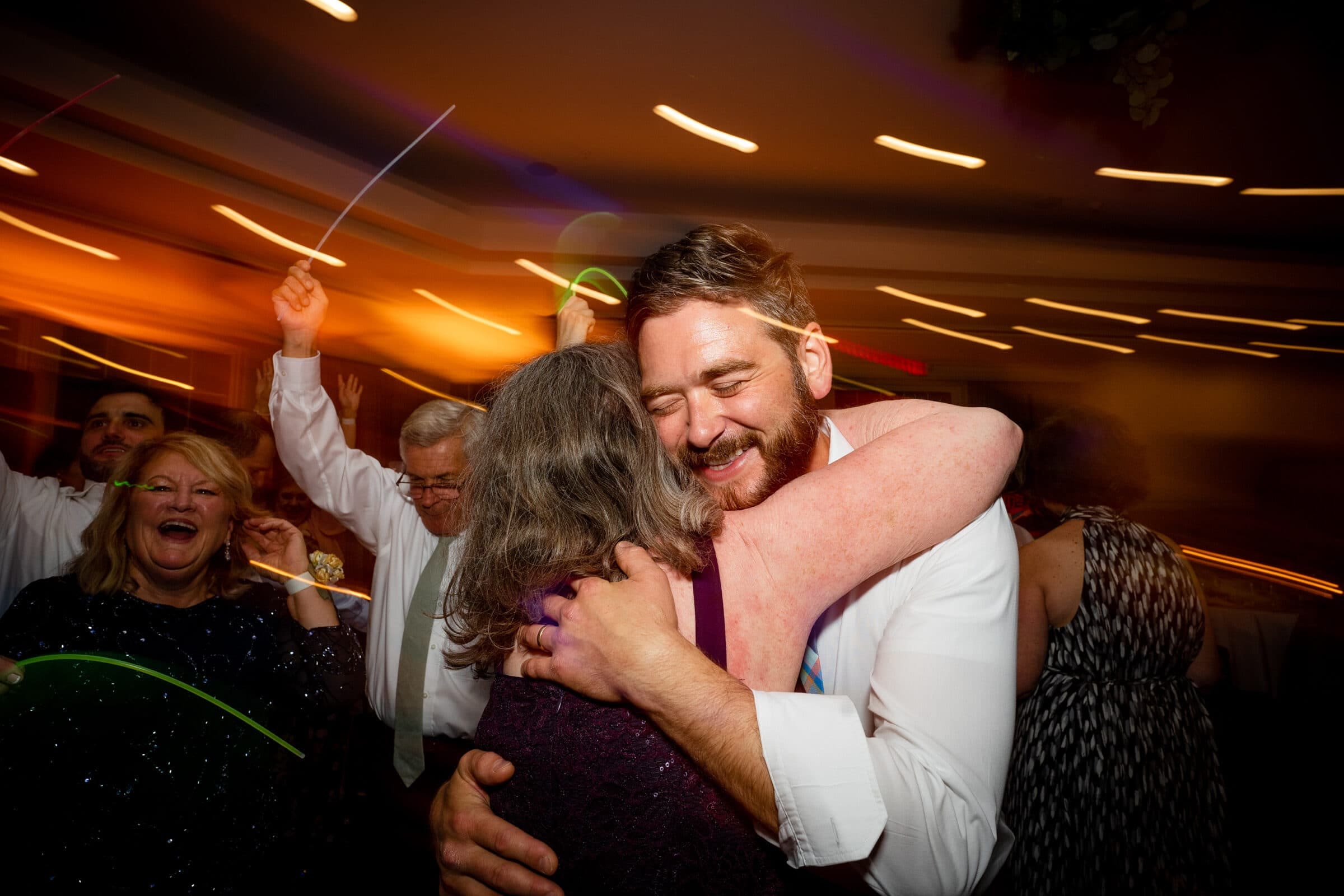 pinehurst wedding photography - lindsey & brendan A joyful scene at a party shows a smiling man embracing a woman. In the background, people are celebrating with colorful glow sticks under warm lighting. The atmosphere is lively and festive.