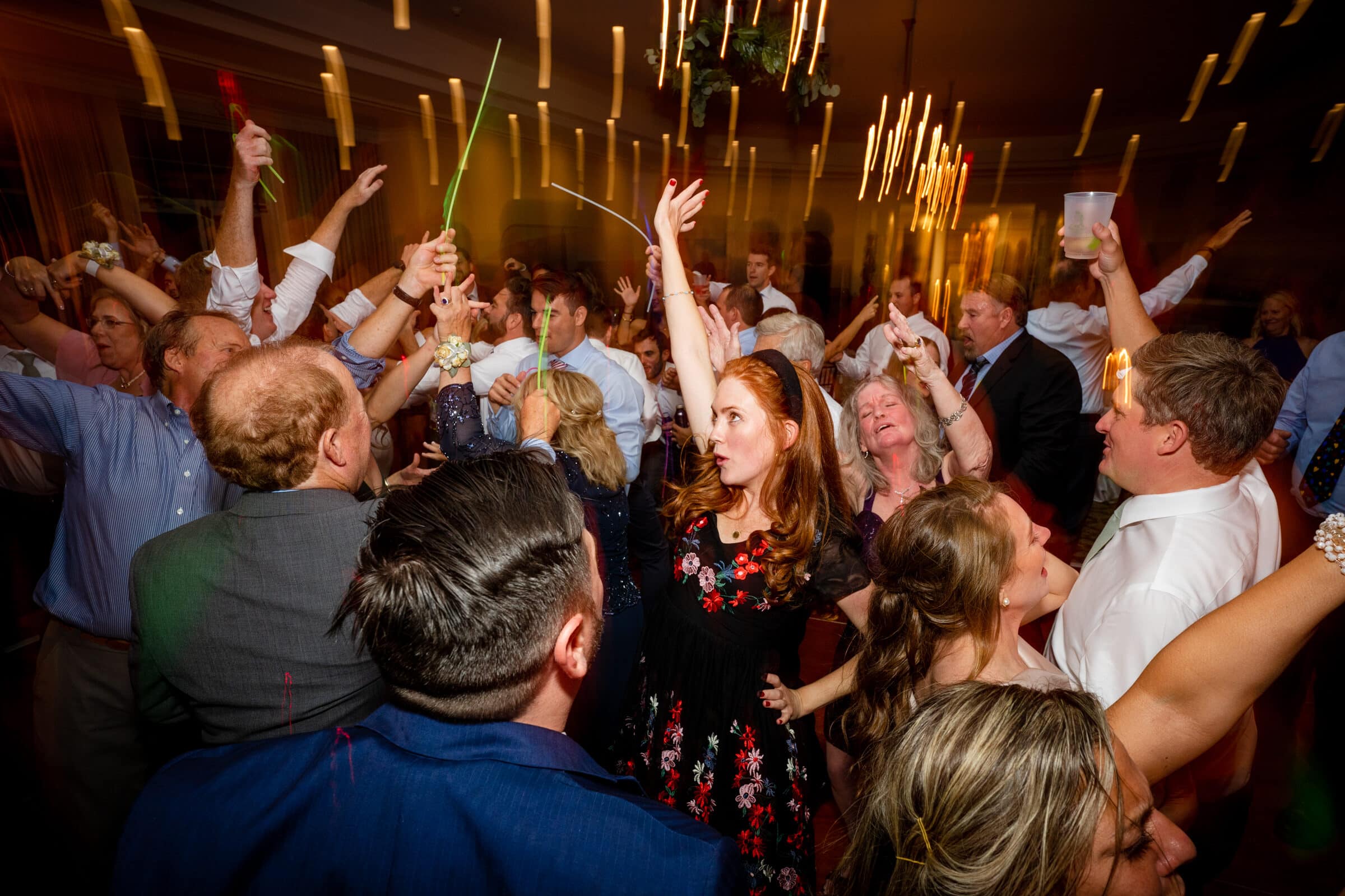 pinehurst wedding photography - lindsey & brendan A lively group of people dancing and celebrating at a party. The room is illuminated with warm, colorful lights, and several guests are raising their hands in the air, creating an energetic atmosphere.