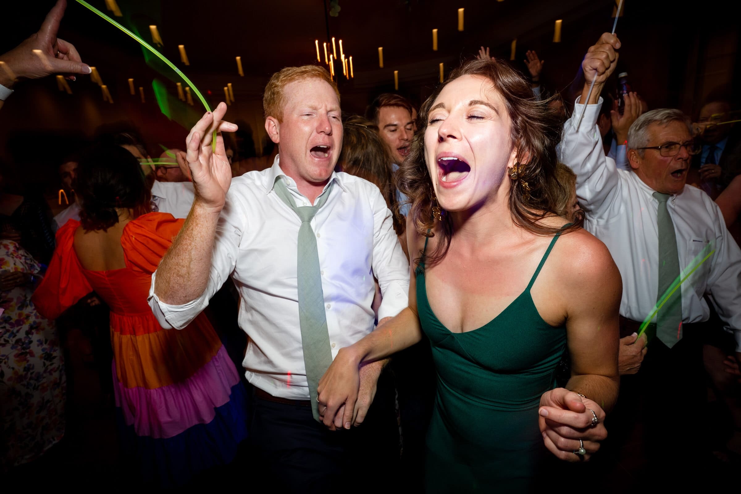 pinehurst wedding photography - lindsey & brendan A lively party scene with people dancing and singing. A man in a white shirt and green tie enthusiastically participates, while a woman in a green dress energetically joins in. The background is filled with colorful lights and partygoers.