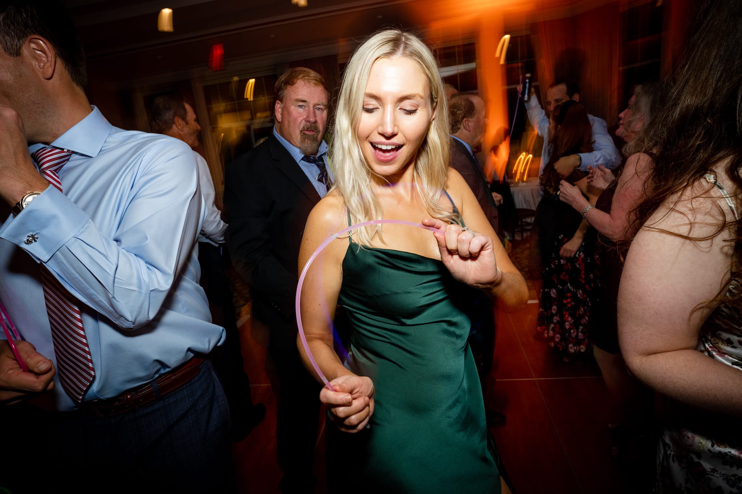 pinehurst wedding photography - lindsey & brendan A woman in a green dress dances energetically at a party, holding a thin glow stick. Around her, other guests are dancing and mingling under dim, colorful lighting. The scene is lively and joyful.
