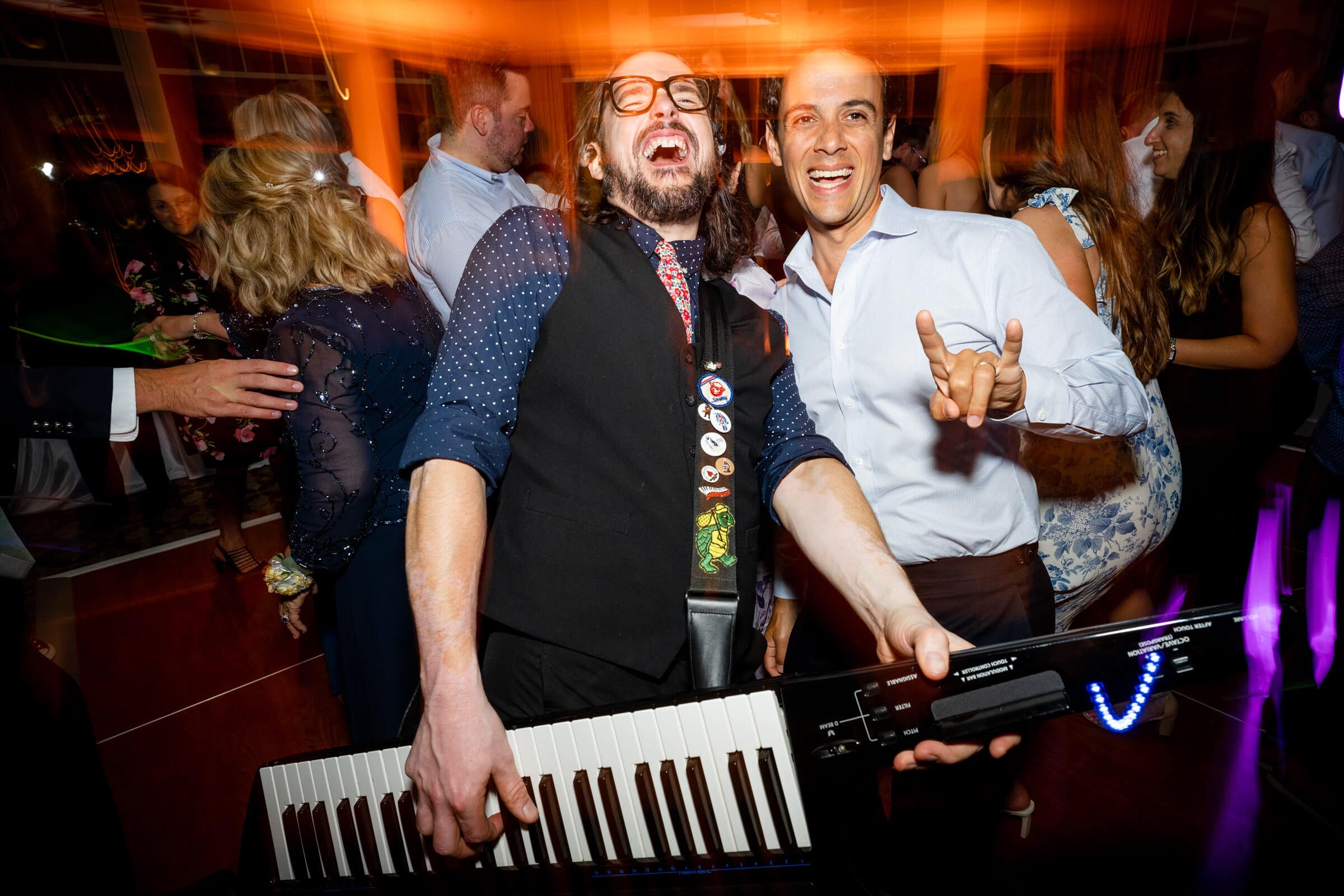 pinehurst wedding photography - lindsey & brendan Two men smiling and posing with a keytar at a lively party. One wears glasses and a vest, holding the keytar with colorful buttons, while the other in a white shirt flashes a rock hand gesture. People in the background are dancing.