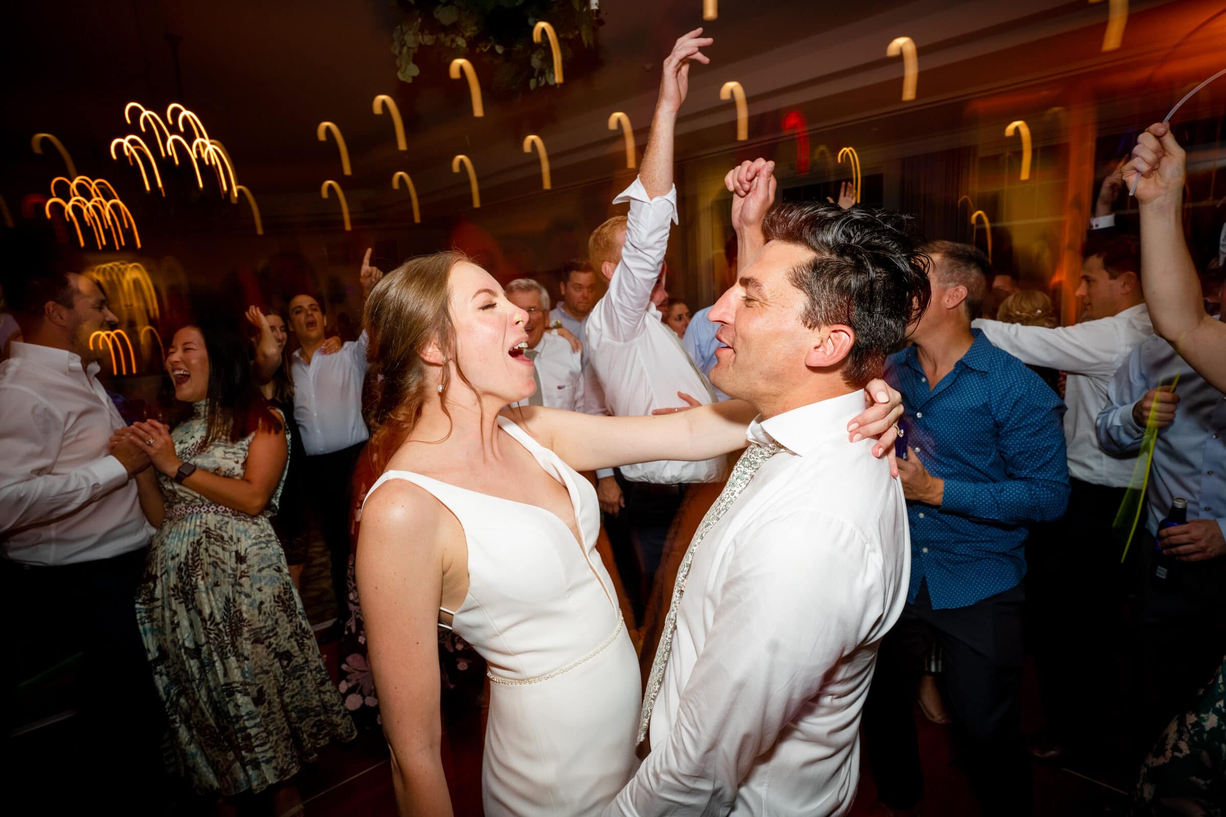 pinehurst wedding photography - lindsey & brendan A joyful couple dances in the center of a lively crowd, surrounded by people clapping and cheering. The woman is in a white dress and the man in a white shirt, with blurred lights creating a festive atmosphere.