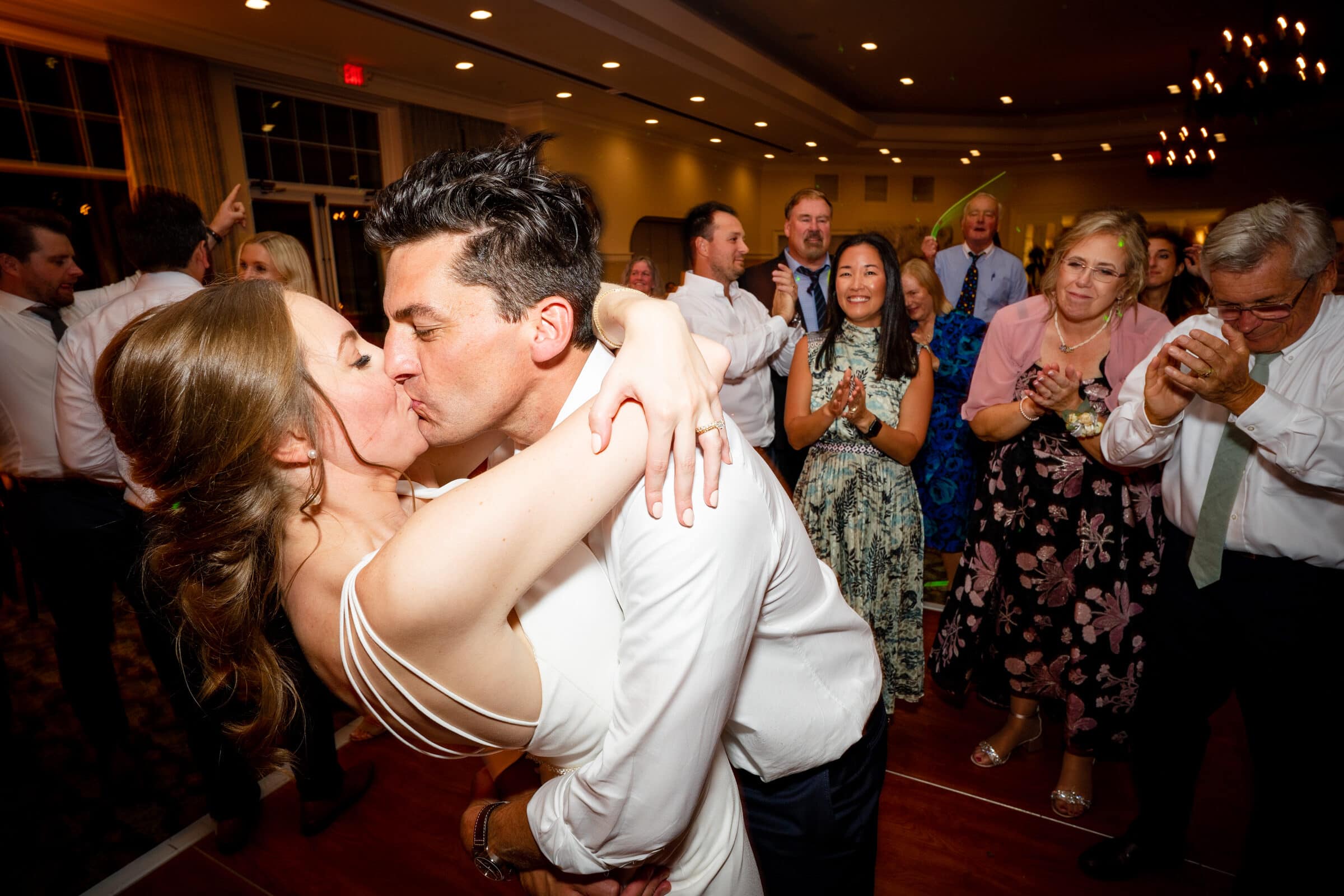 pinehurst wedding photography - lindsey & brendan A newlywed couple shares a romantic kiss on the dance floor, surrounded by cheering and smiling guests in formal attire. The room is warmly lit, creating a joyful and celebratory atmosphere.