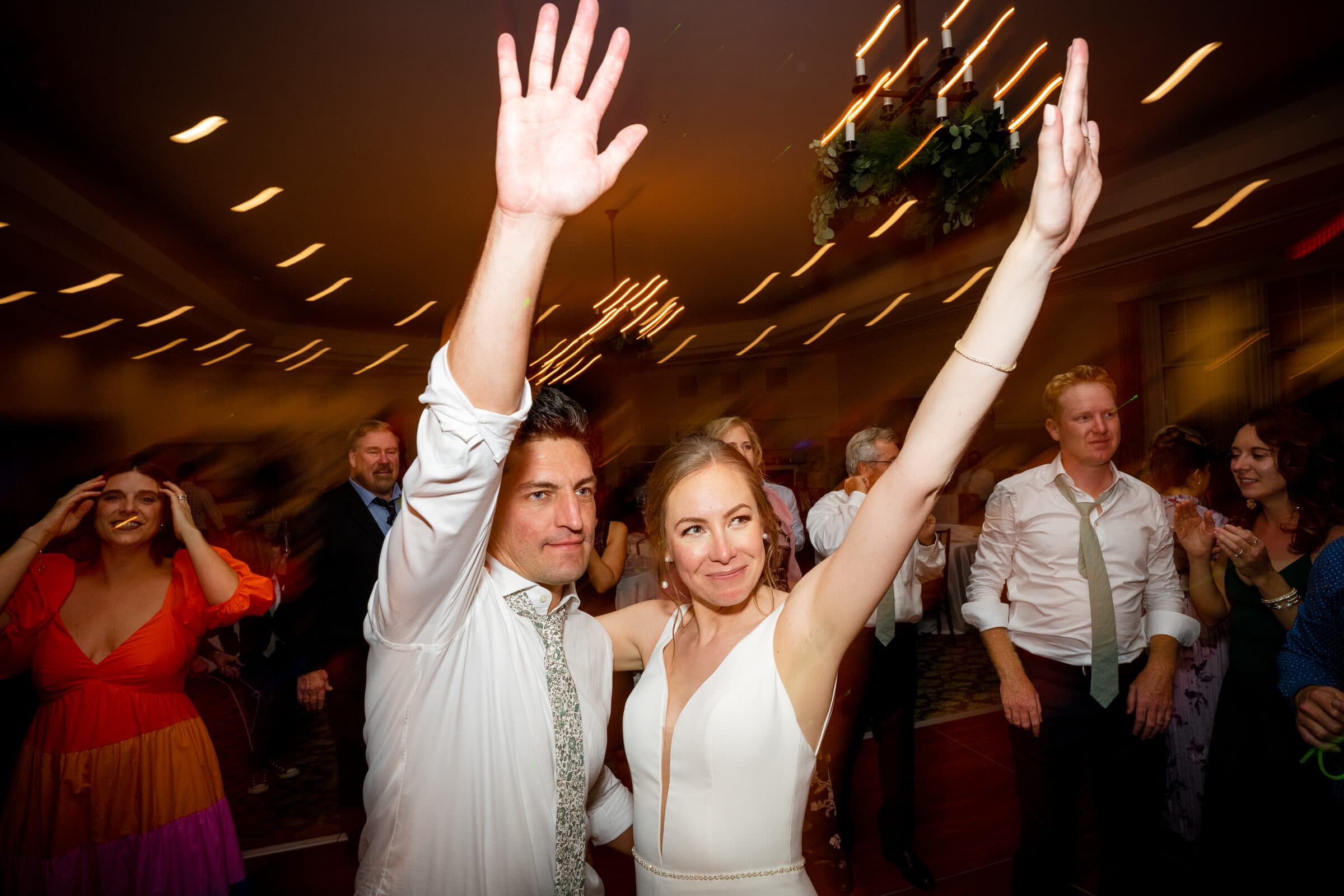 pinehurst wedding photography - lindsey & brendan A couple in formal attire smiles and raises their hands in celebration on a dance floor surrounded by friends. The room is warmly lit with chandeliers, and people around them are enjoying the festive atmosphere.