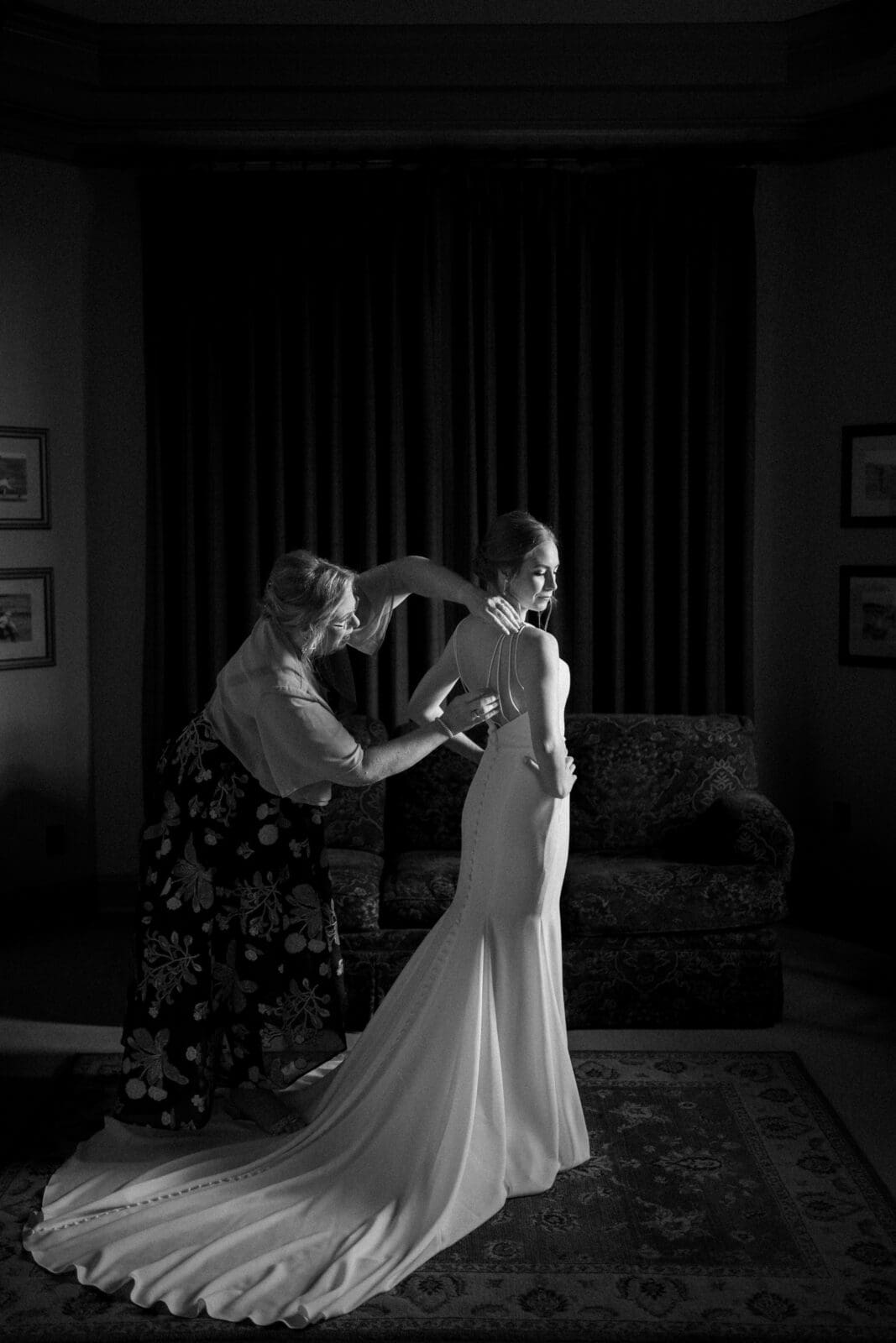 A woman assists a bride with her wedding dress. The bride stands in a dimly lit room, wearing a long, elegant gown with a train. The ambiance is intimate, with soft lighting highlighting the intricate dress details.