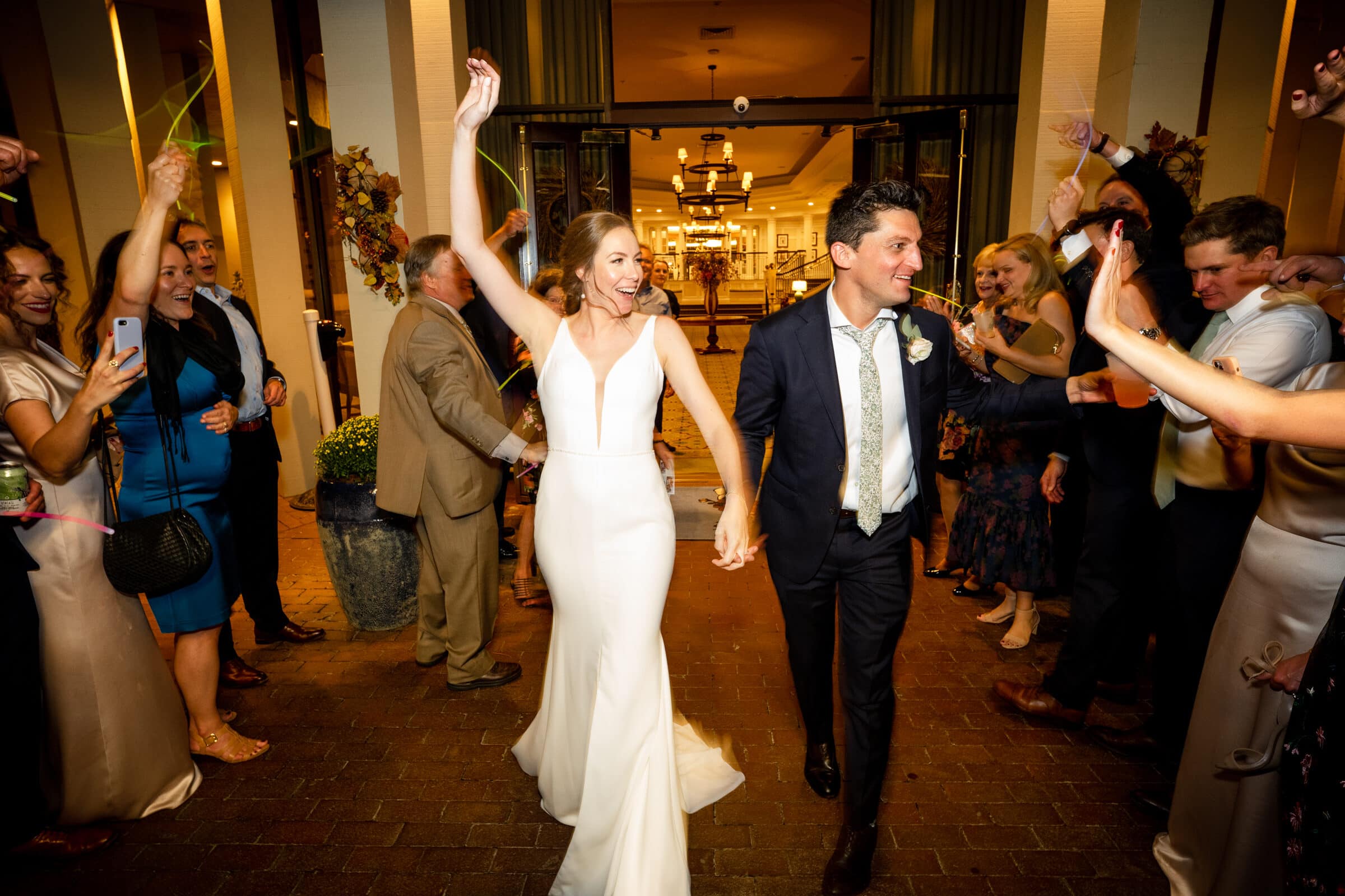 A joyful bride in a white dress and a groom in a suit walk through a cheering crowd celebrating their wedding. People around them hold streamers and take photos, creating a festive atmosphere.