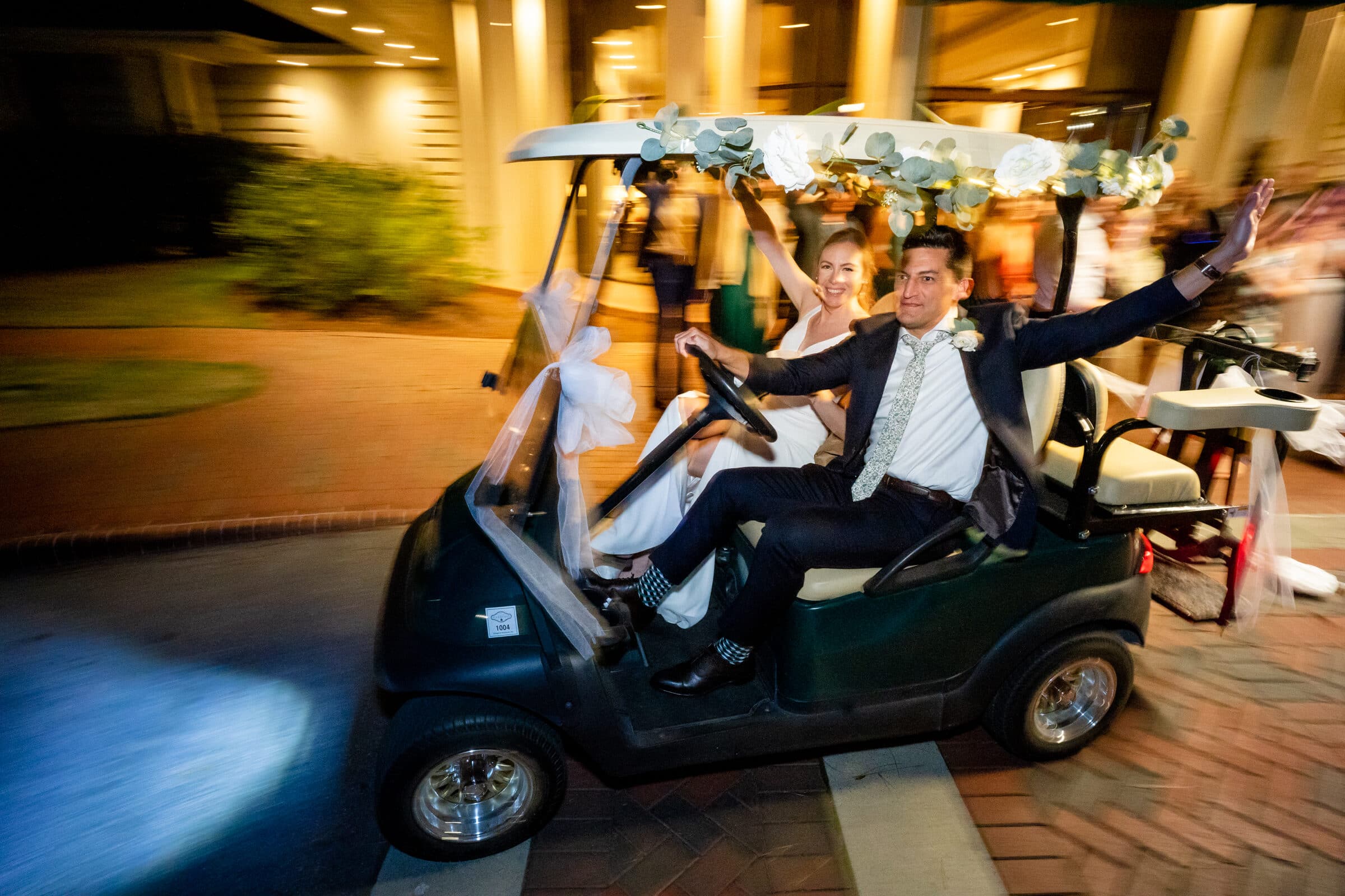 pinehurst wedding photography - lindsey & brendan A joyful couple wearing formal attire rides in a decorated golf cart. The groom raises his hands in excitement, while the bride smiles beside him. The background shows a building with onlookers and blurred lights, suggesting a festive occasion.