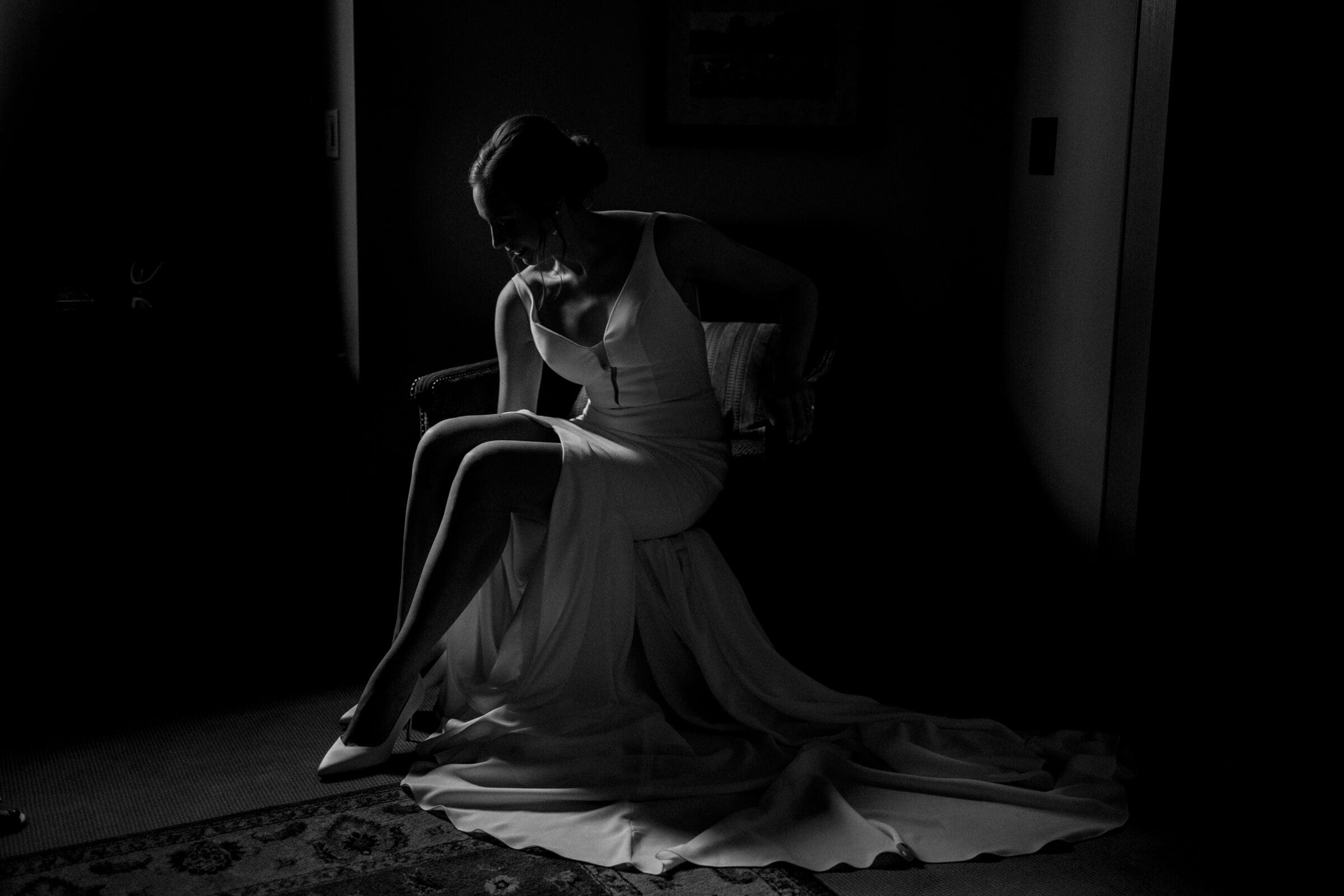 pinehurst wedding photography - lindsey & brendan A woman in a long, elegant gown sits on a chair in a dimly lit room. Her head is bowed slightly, and her dress flows onto the floor, illuminated by light streaming from the side. The mood is calm and contemplative.