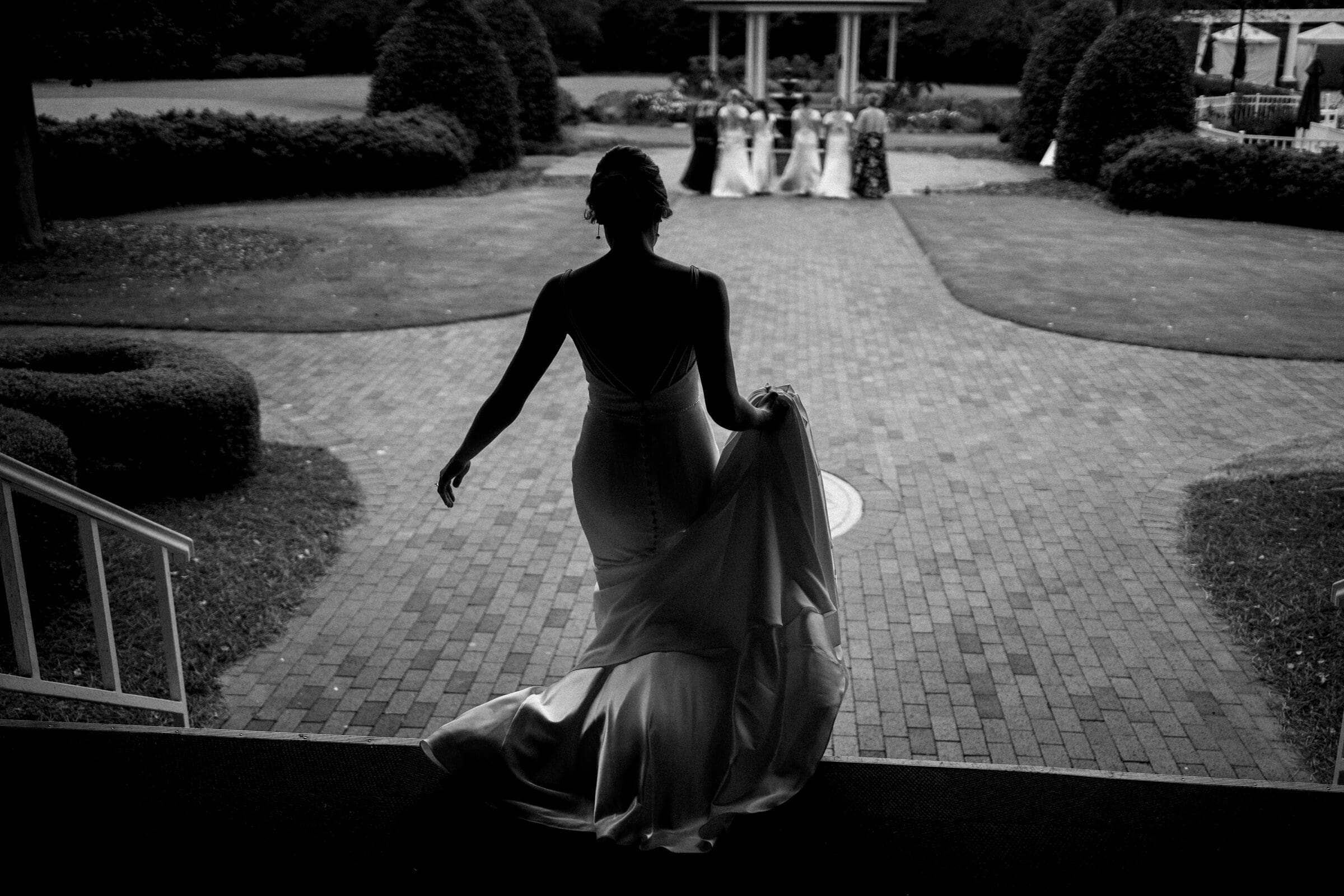 pinehurst wedding photography - lindsey & brendan A silhouetted bride in a flowing gown descends steps towards a garden path, where a wedding ceremony is taking place. Bridesmaids and groomsmen are visible in the distance, standing under a gazebo amidst lush greenery.