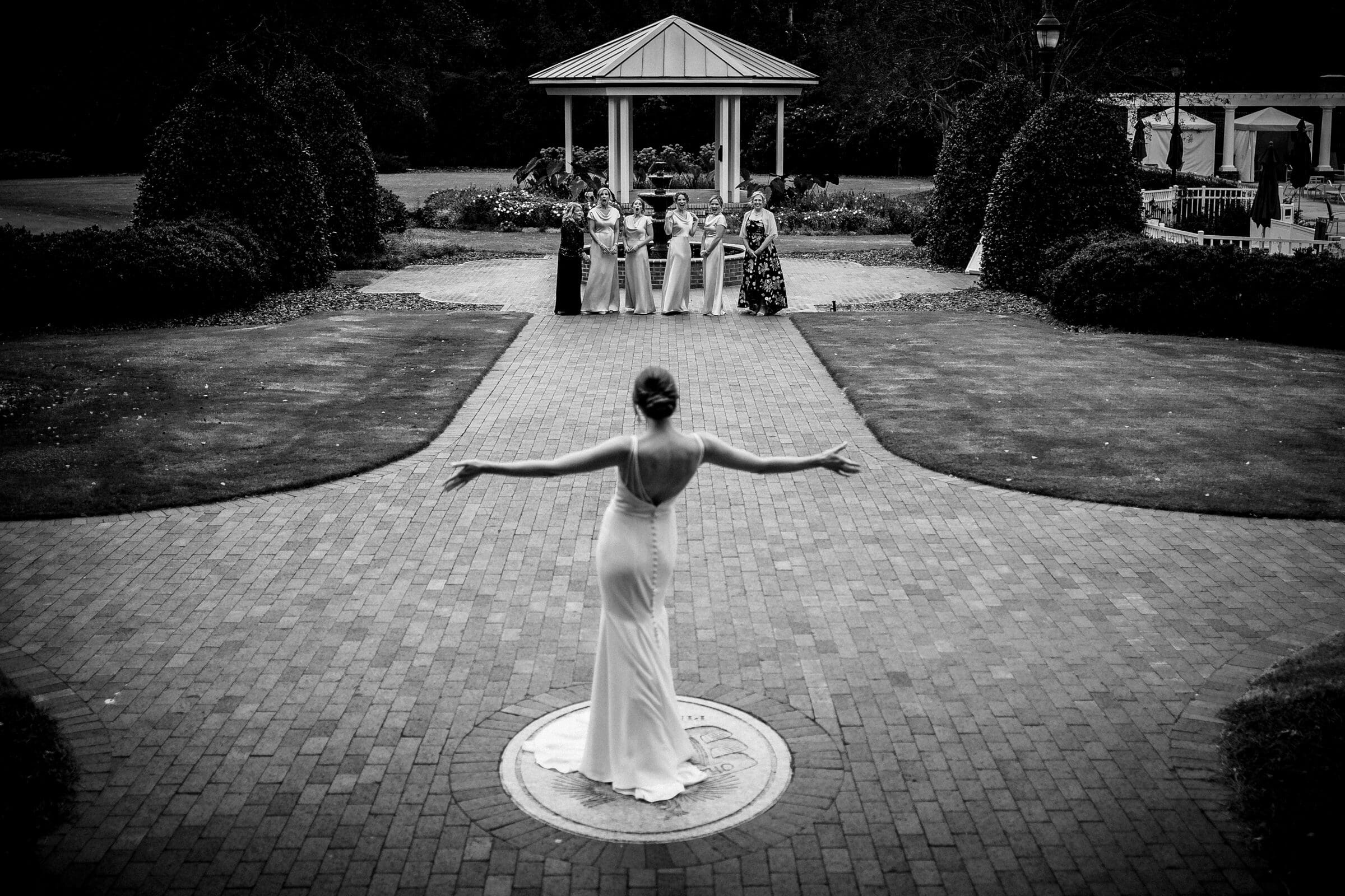A person in a white gown stands with arms outstretched on a brick path, facing away. In the background, a group of people stand near a gazebo, watching. The setting is an outdoor garden with neatly trimmed hedges. Black and white photograph.