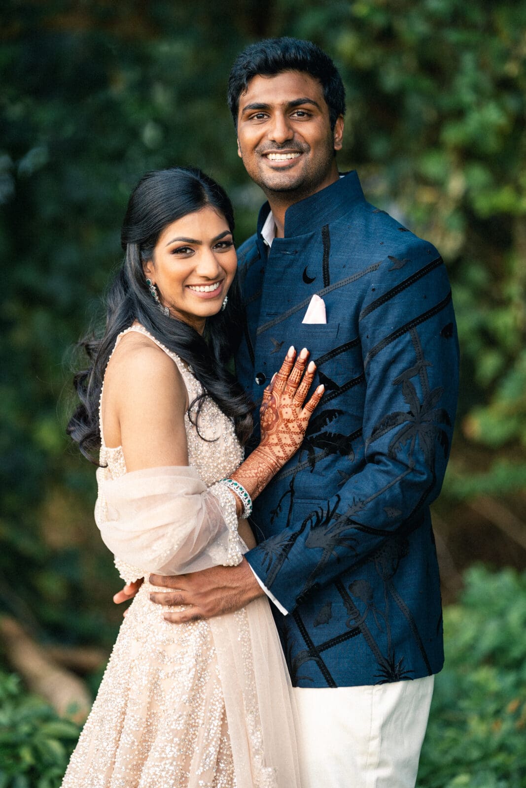A couple smiles warmly at the camera in an outdoor setting. The woman wears a light-colored embellished gown with henna on her hands, and the man is dressed in a blue jacket and white pants. They are surrounded by lush greenery.