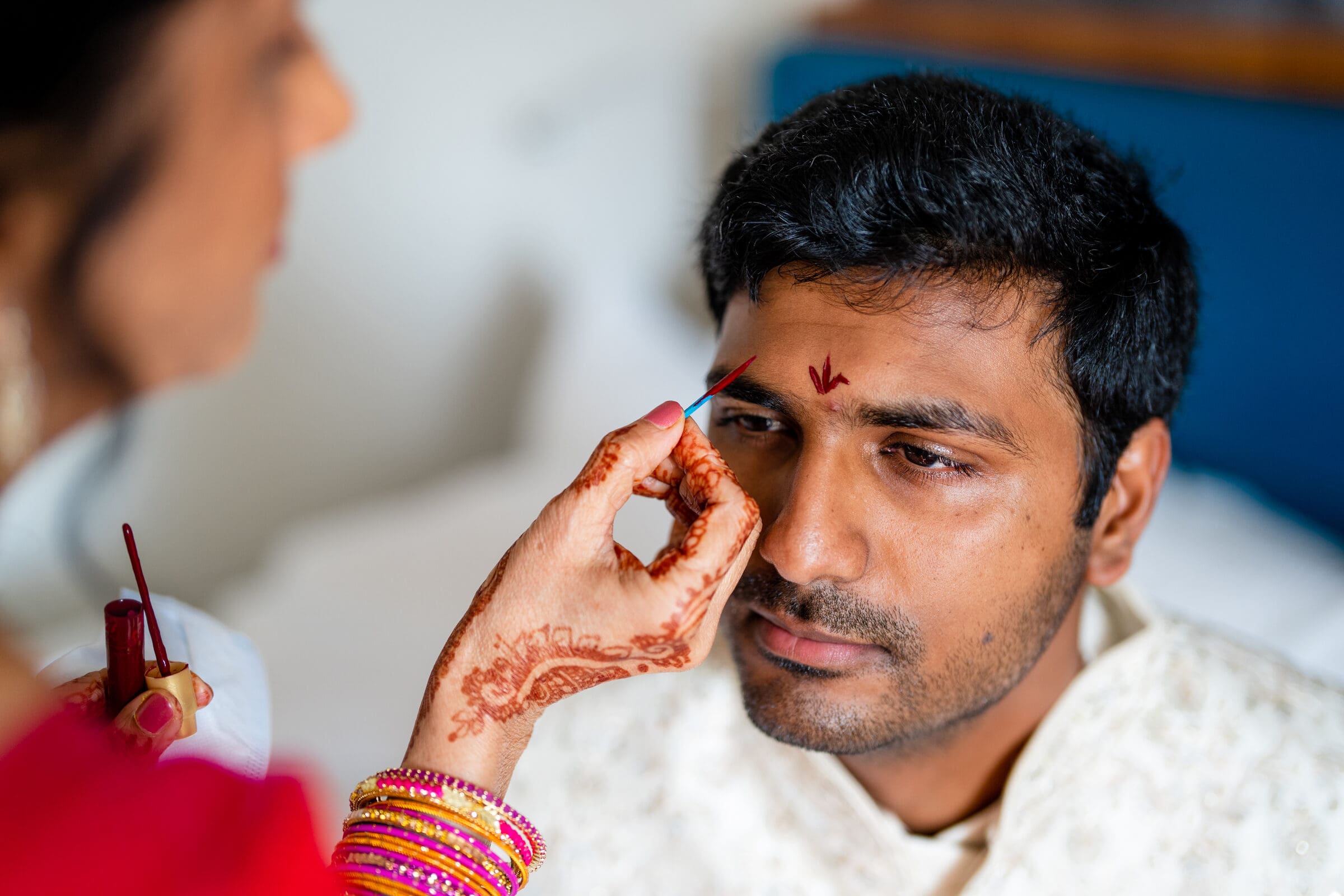 A person with mehndi on their hand applies a red mark on another persons forehead, who is wearing a white traditional outfit. The scene suggests a cultural or religious ceremony.