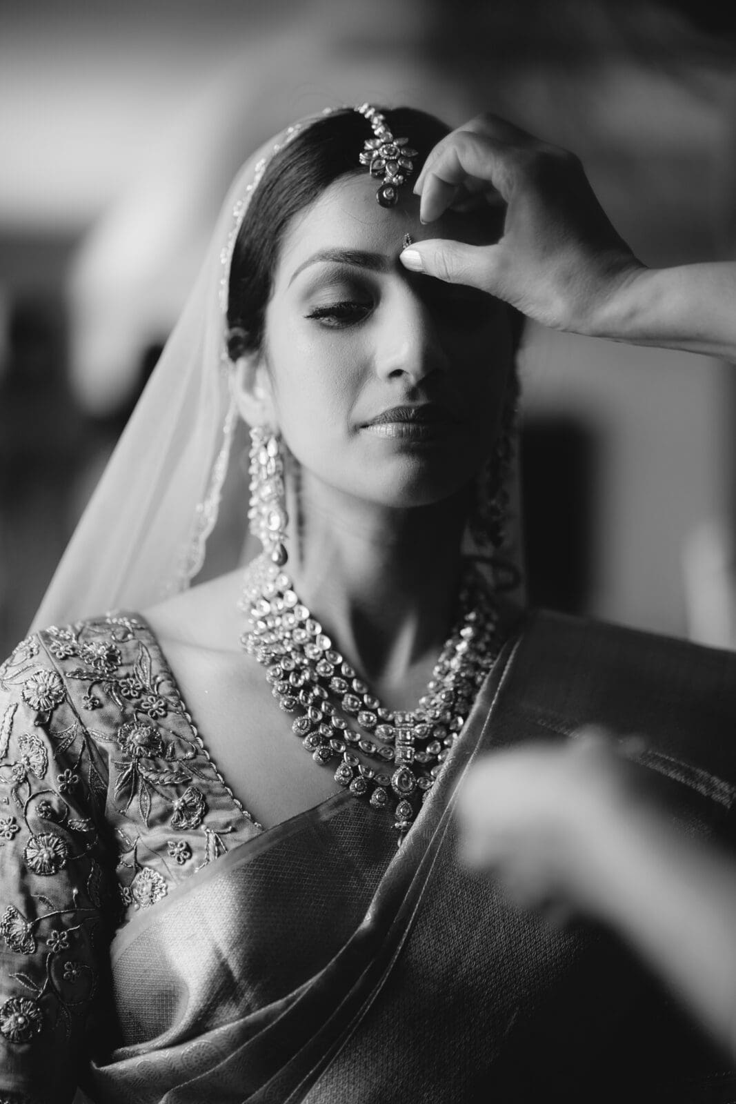 A bride in traditional attire with intricate jewelry closes her eyes as someone gently adjusts her bindi. She wears an embroidered saree, and the scene is captured in elegant black and white.