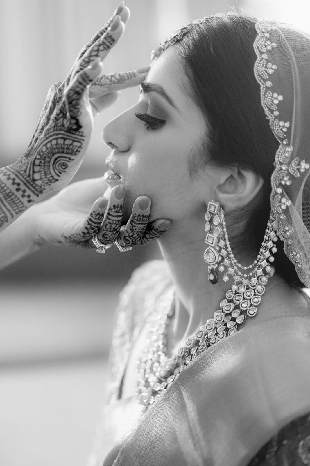 A black and white photo of a bride getting ready. The bride has intricate henna designs on her hands, wears ornate jewelry, including a necklace and earrings, and a veil. A hand with henna applies a bindi to her forehead. The scene is serene and elegant.