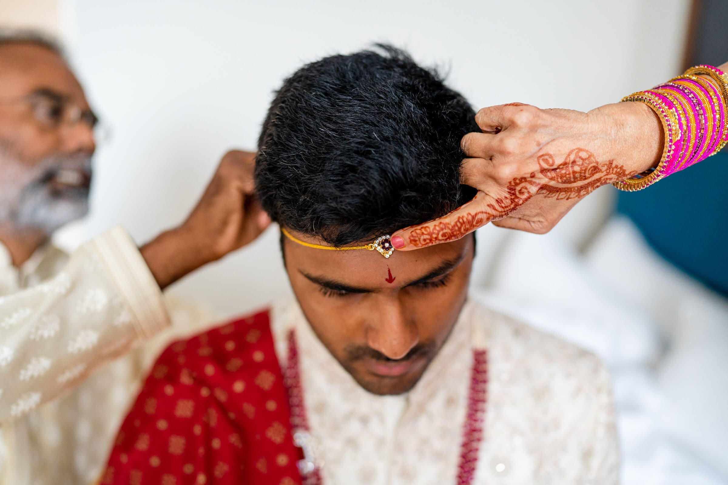 A person carefully adjusts a golden headband on a mans head, who is seated with eyes closed. The mans forehead is adorned with a red symbol, and hes wearing a decorated outfit. Another person assists from the side, dressed in a light-colored garment.