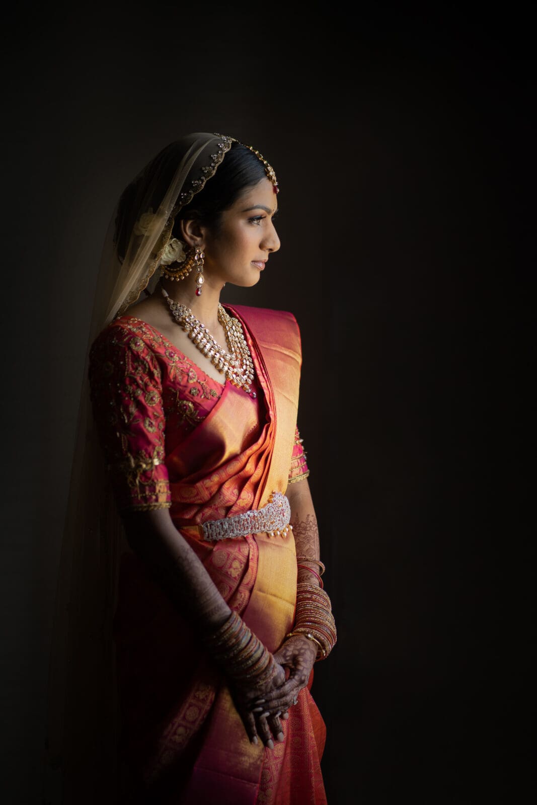 A bride in a red and gold saree stands against a dark background. She is adorned with traditional jewelry and her hands display intricate henna designs. The bride gazes thoughtfully into the distance.