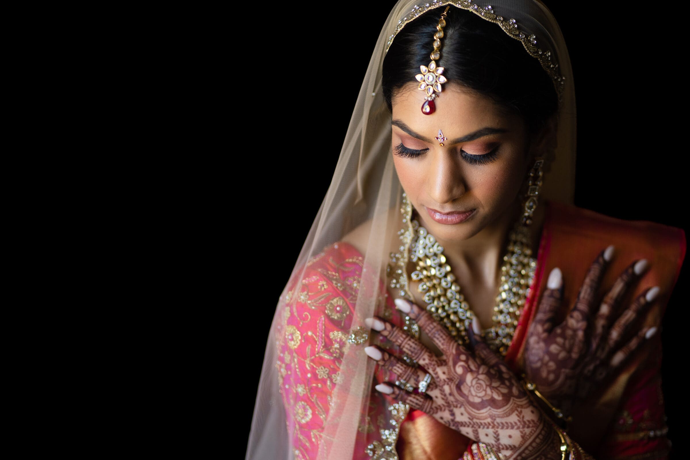 A bride in a traditional Indian outfit, featuring intricate henna on her hands, adorned with jewelry and a veil. She holds her hands gracefully against her chest, exuding elegance and poise against a black background.