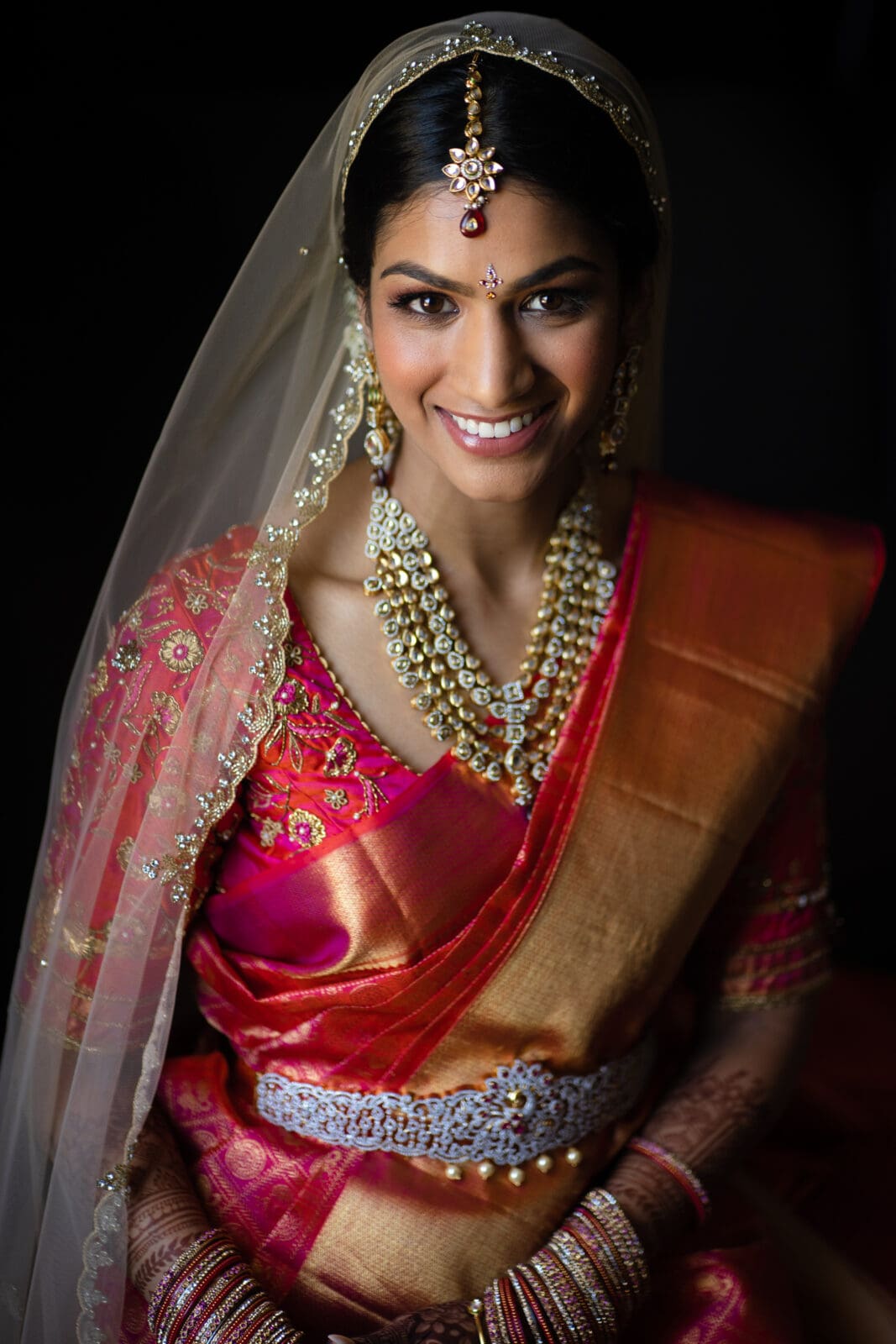 A smiling bride in a traditional Indian outfit with a red and gold saree, adorned with intricate jewelry, including a necklace and headpiece. Her hands are decorated with henna, and she wears a sheer veil.