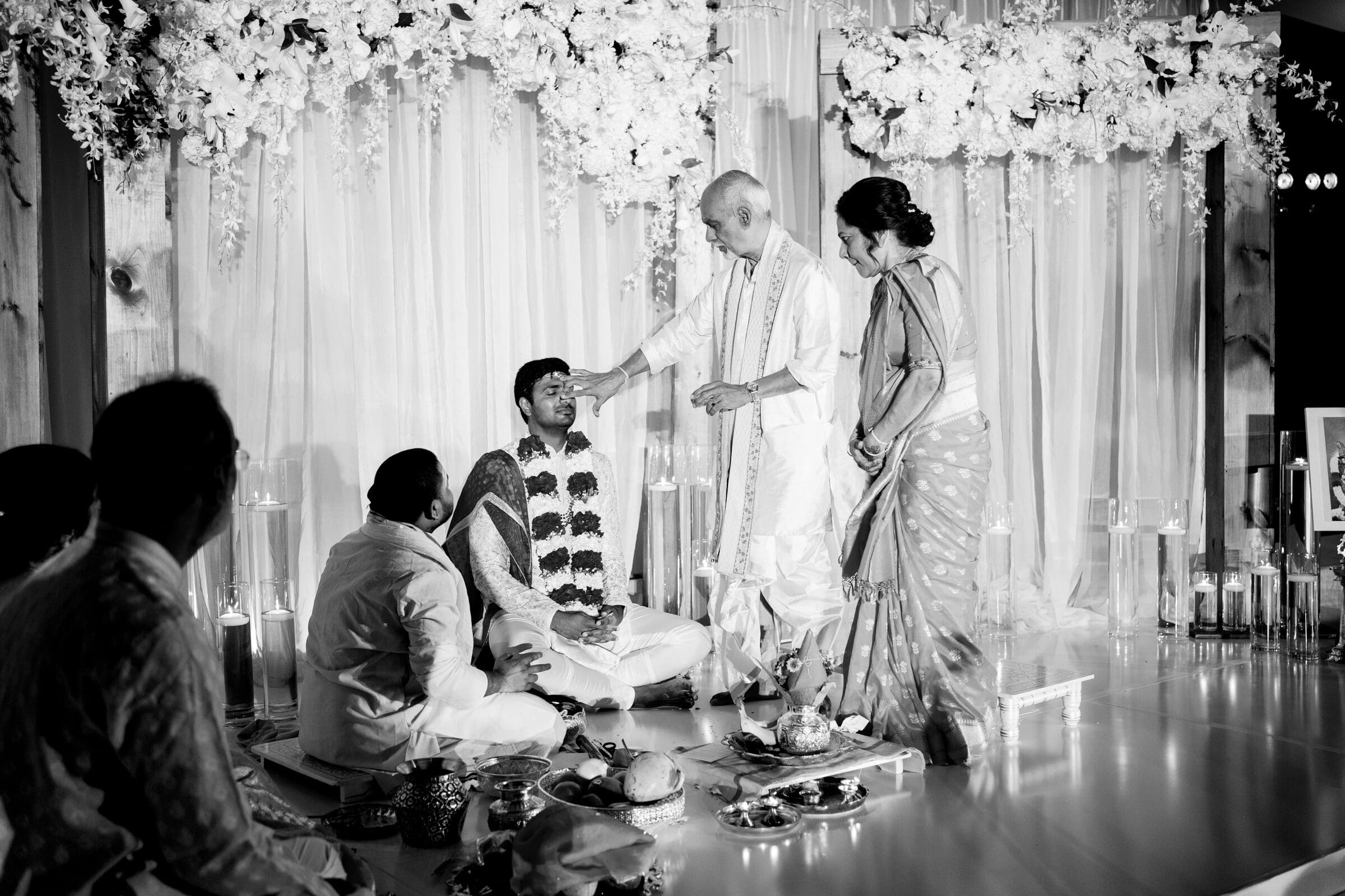 A black and white image shows a couple engaging in a ritual with a man, possibly during a wedding ceremony. They stand on a decorated stage, surrounded by floral arrangements. Seated individuals observe, and traditional items are on the floor.