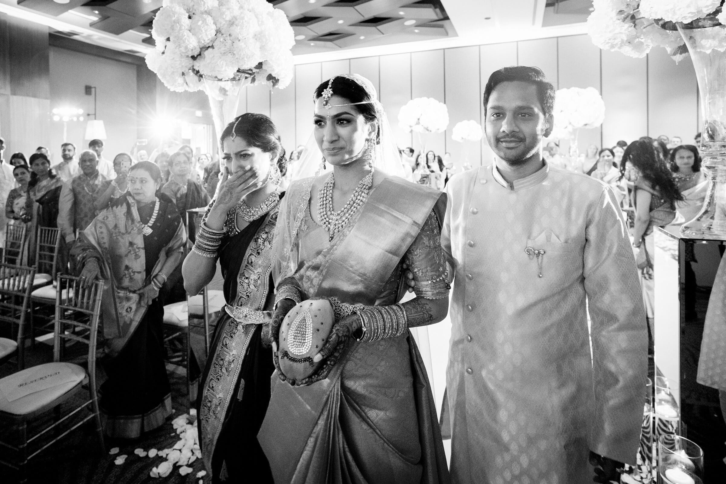 A bride in a traditional sari, accompanied by two people, walks down an aisle during a wedding ceremony. The scene is indoors with many guests and floral decorations. The atmosphere is solemn and elegant.