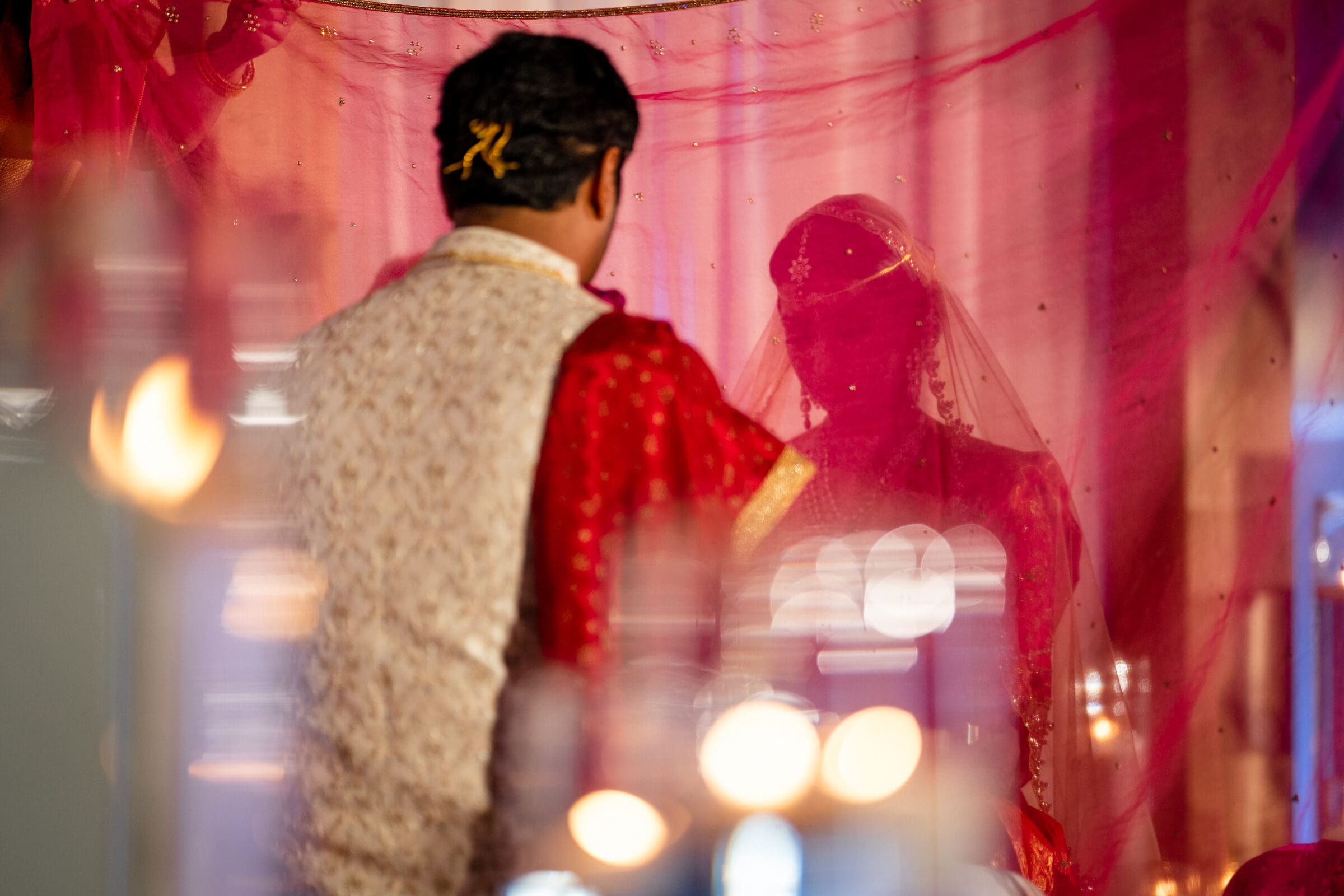 A couple in traditional attire stands in front of a pink backdrop. The groom faces away, wearing a gold and red outfit, while the bride, adorned with jewelry, appears partially obscured by soft lighting and a veil.