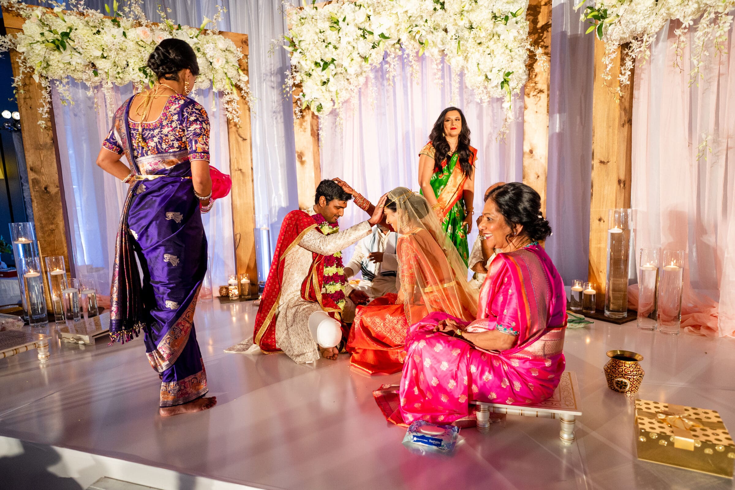 A couple in traditional attire participating in a wedding ceremony. The groom places a garland on the bride. Two women in colorful saris sit nearby, while another woman stands in the background. The setting is decorated with flowers and candles.