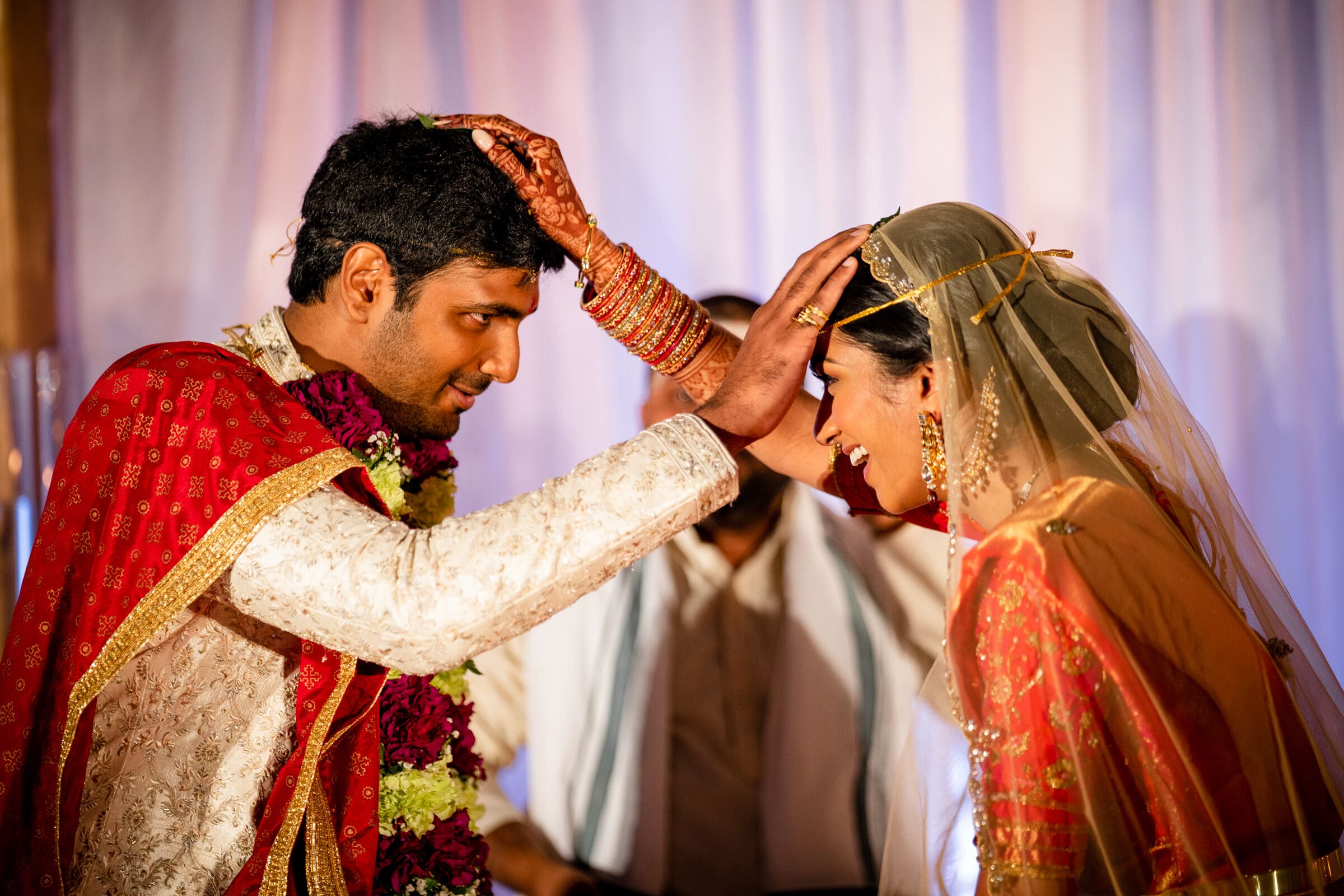 A couple in traditional wedding attire exchanges garlands during a ceremony. The bride wears an ornate red veil and jewelry, while the groom is dressed in a matching embroidered outfit. Both are smiling, immersed in the ritual.