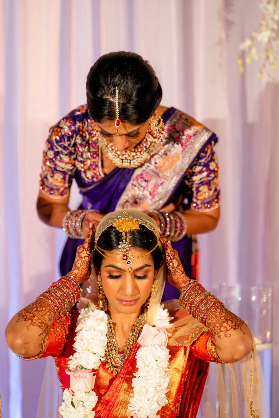 A woman in a red and gold sari, adorned with henna and jewelry, sits while another woman in a purple sari adjusts her headpiece. The scene is set against a soft curtain backdrop, with floral decorations partly visible.